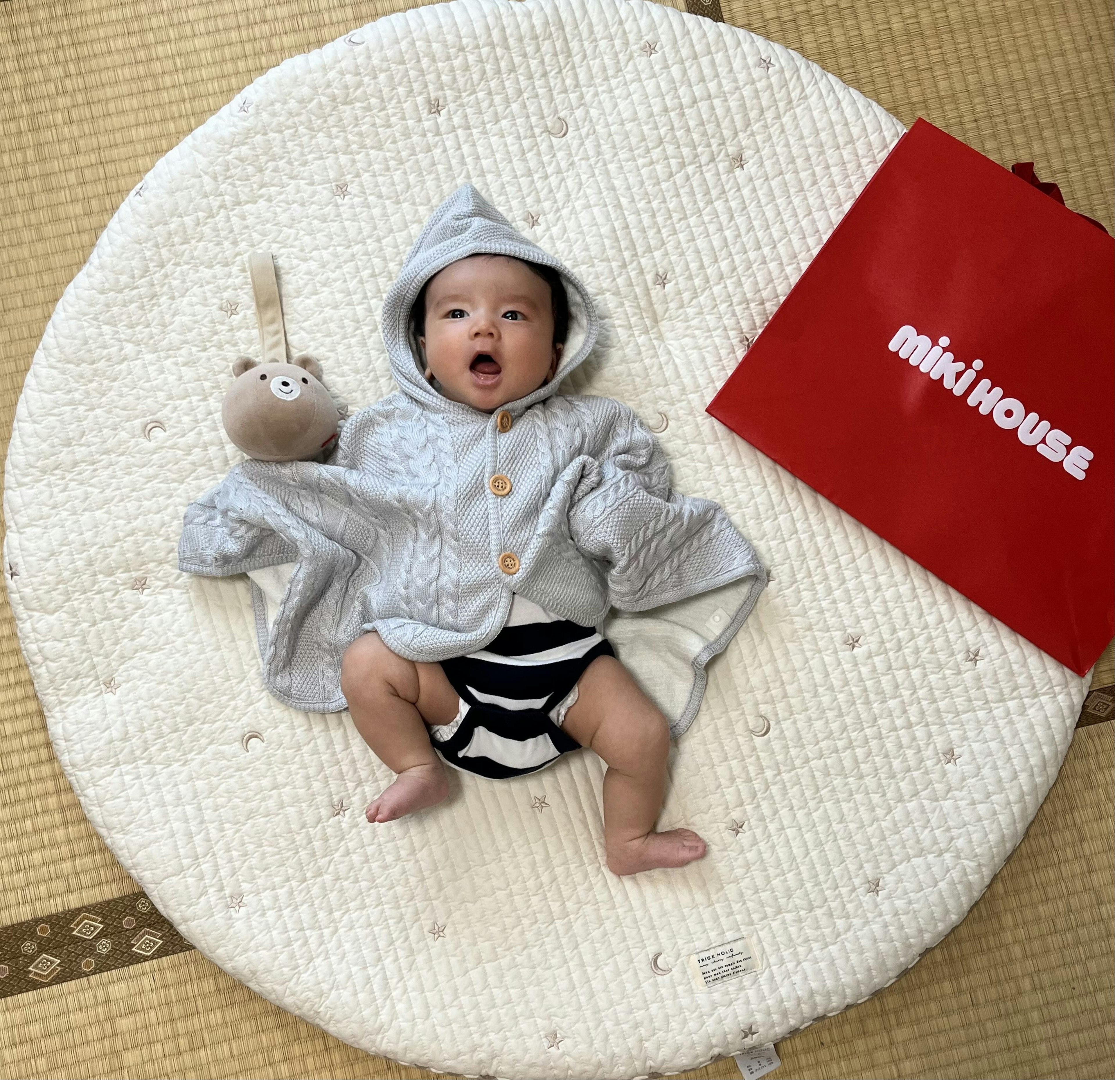 Bebé en un atuendo gris con capucha sosteniendo un juguete de peluche junto a una bolsa roja de miki house