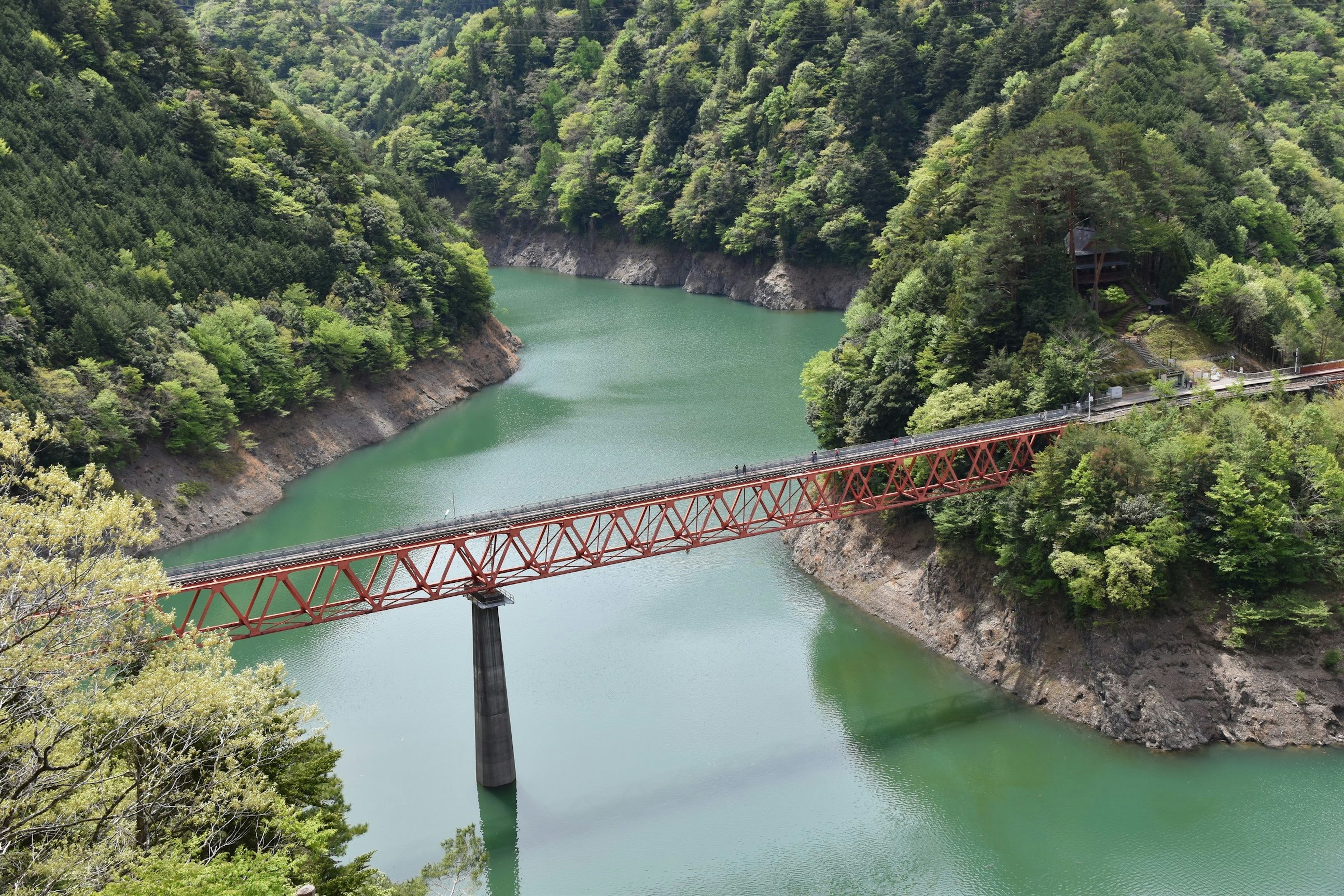 Jembatan besi merah di atas air hijau dikelilingi gunung subur