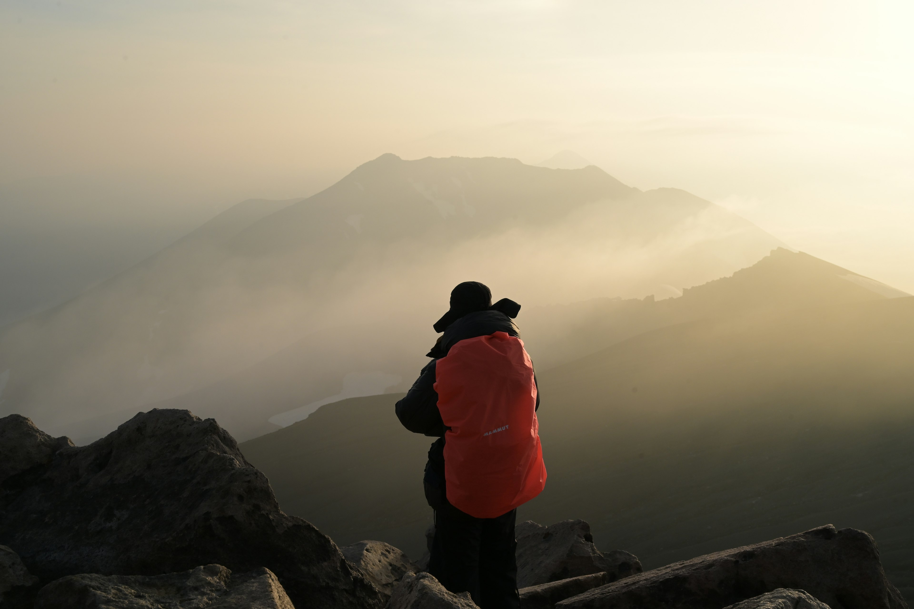 山頂でオレンジのバックパックを背負った登山者が霧に包まれた山々を眺めている