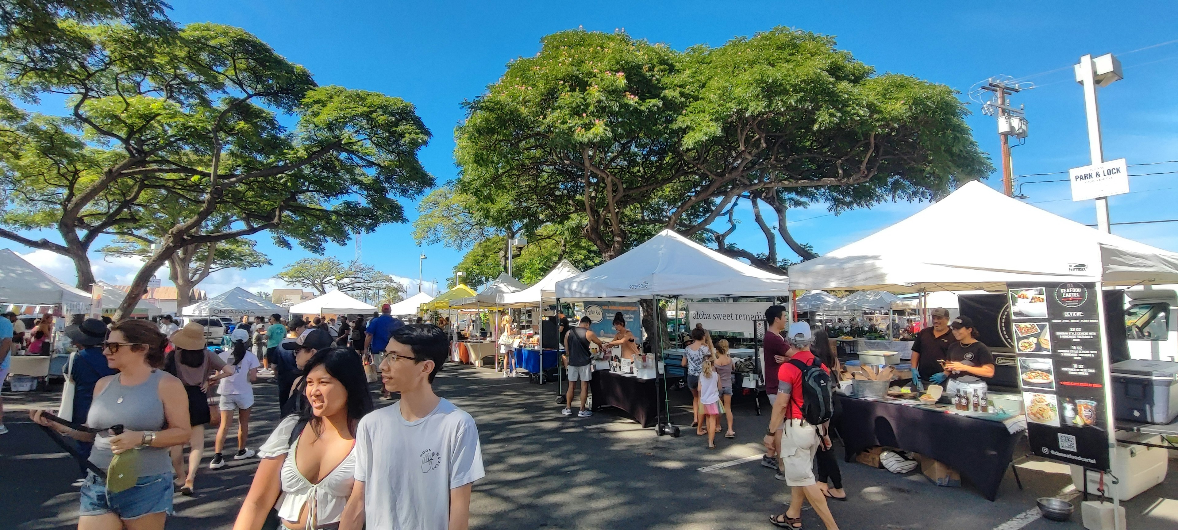 A bustling street market scene under a blue sky featuring many people and white tents