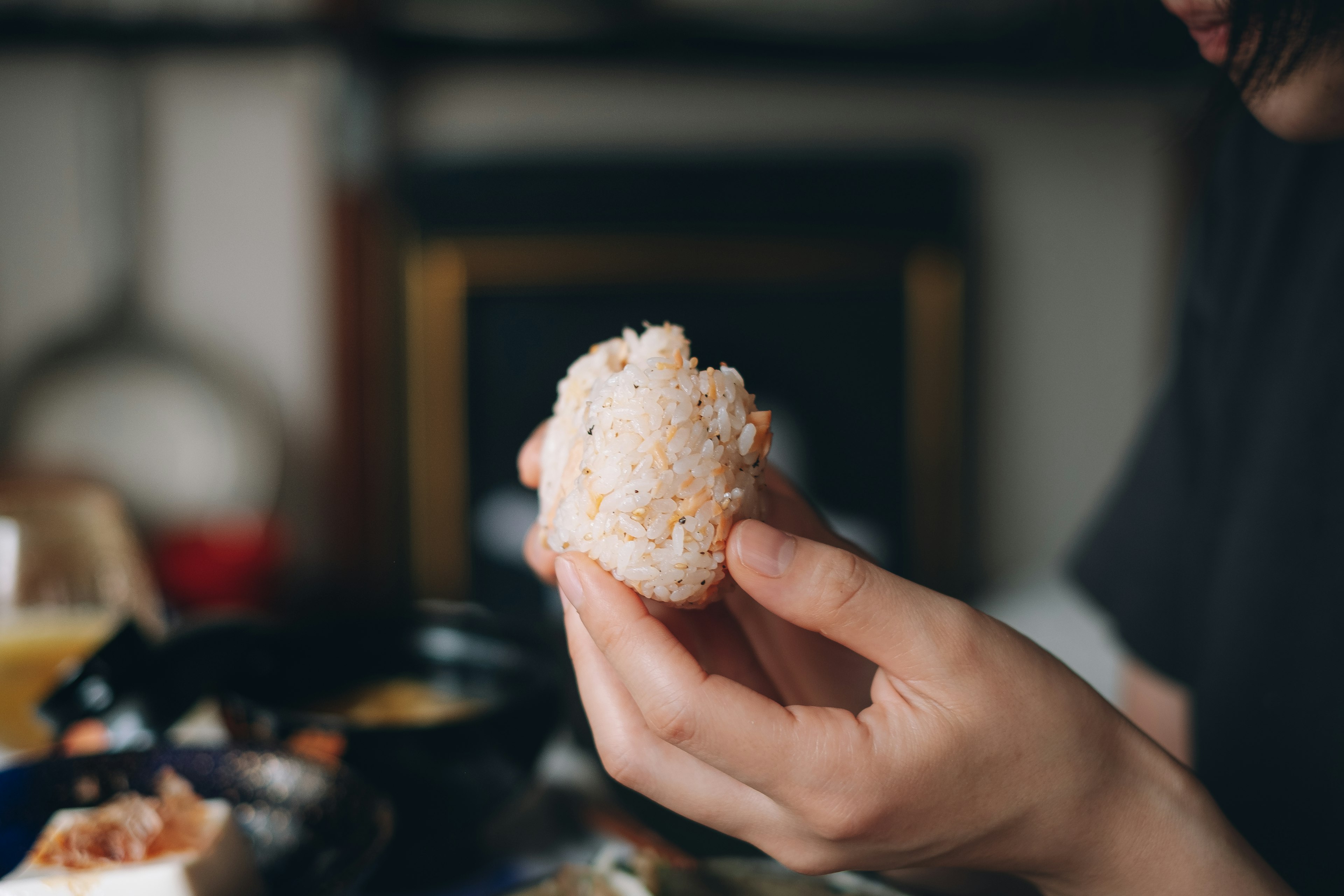 A hand holding a rice ball with various dishes in the background