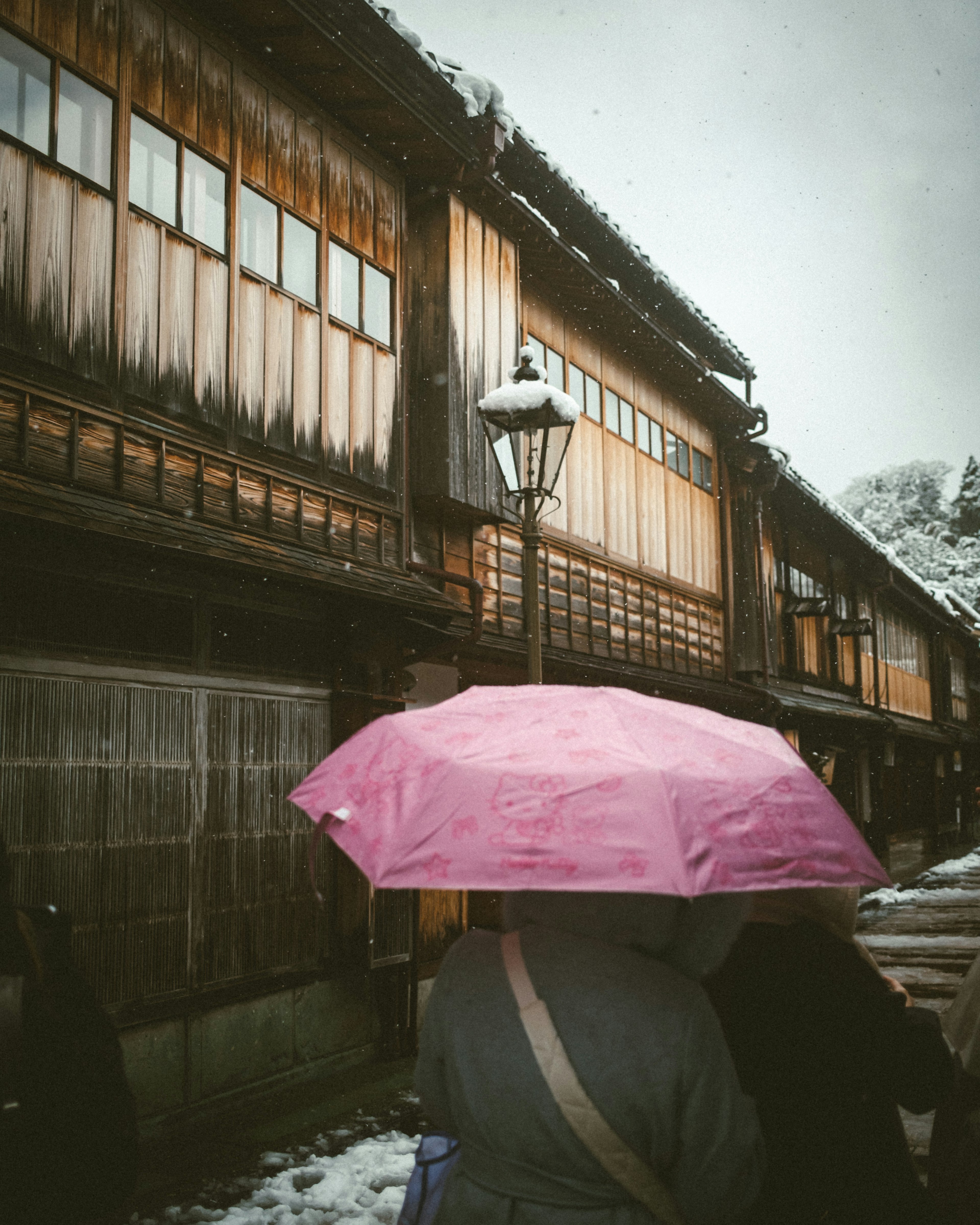 雪の降る中、ピンクの傘を持った人が古い木造の建物の前を歩いている情景