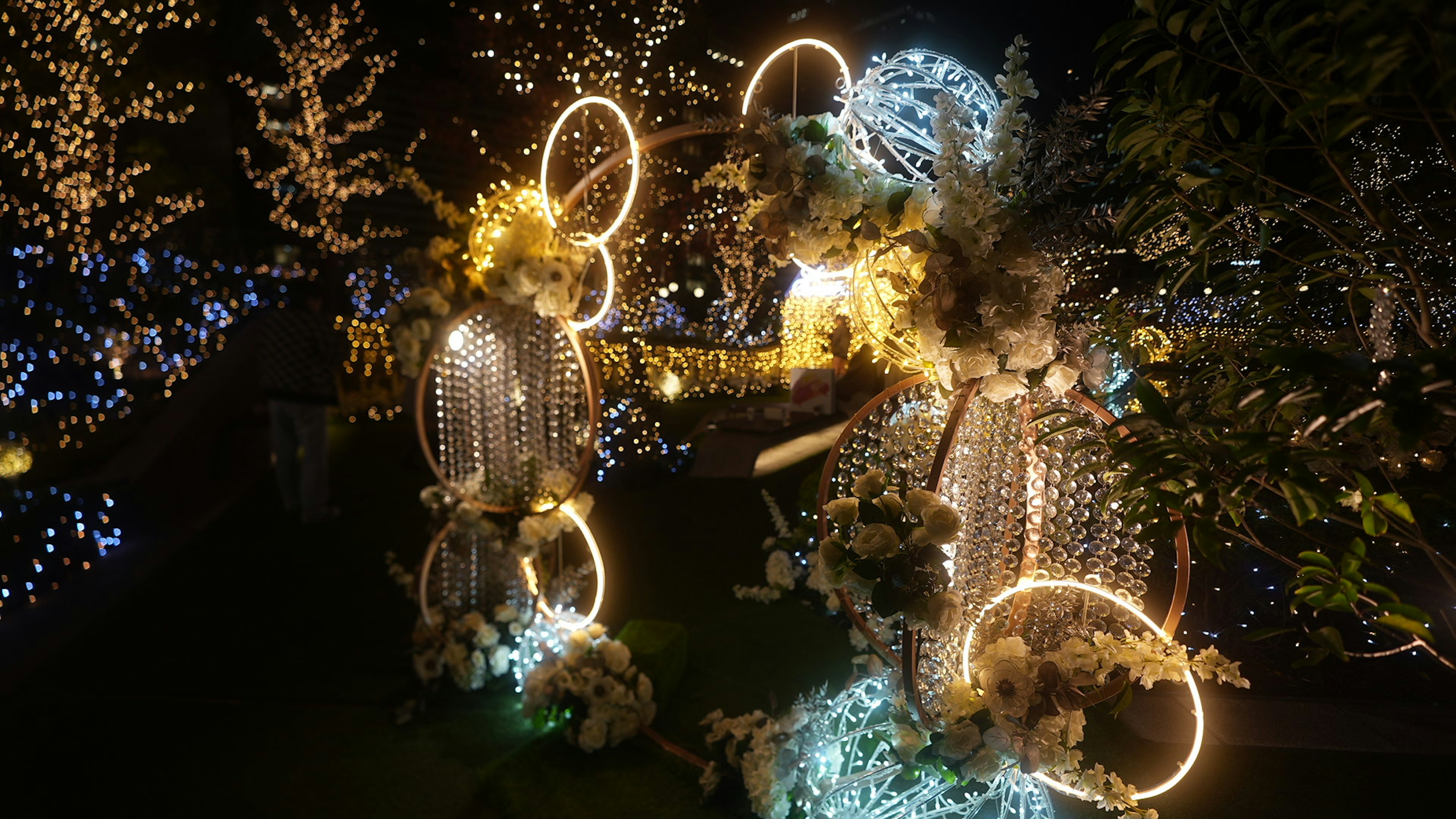 Two illuminated figures adorned with flowers and lights in a nighttime garden setting