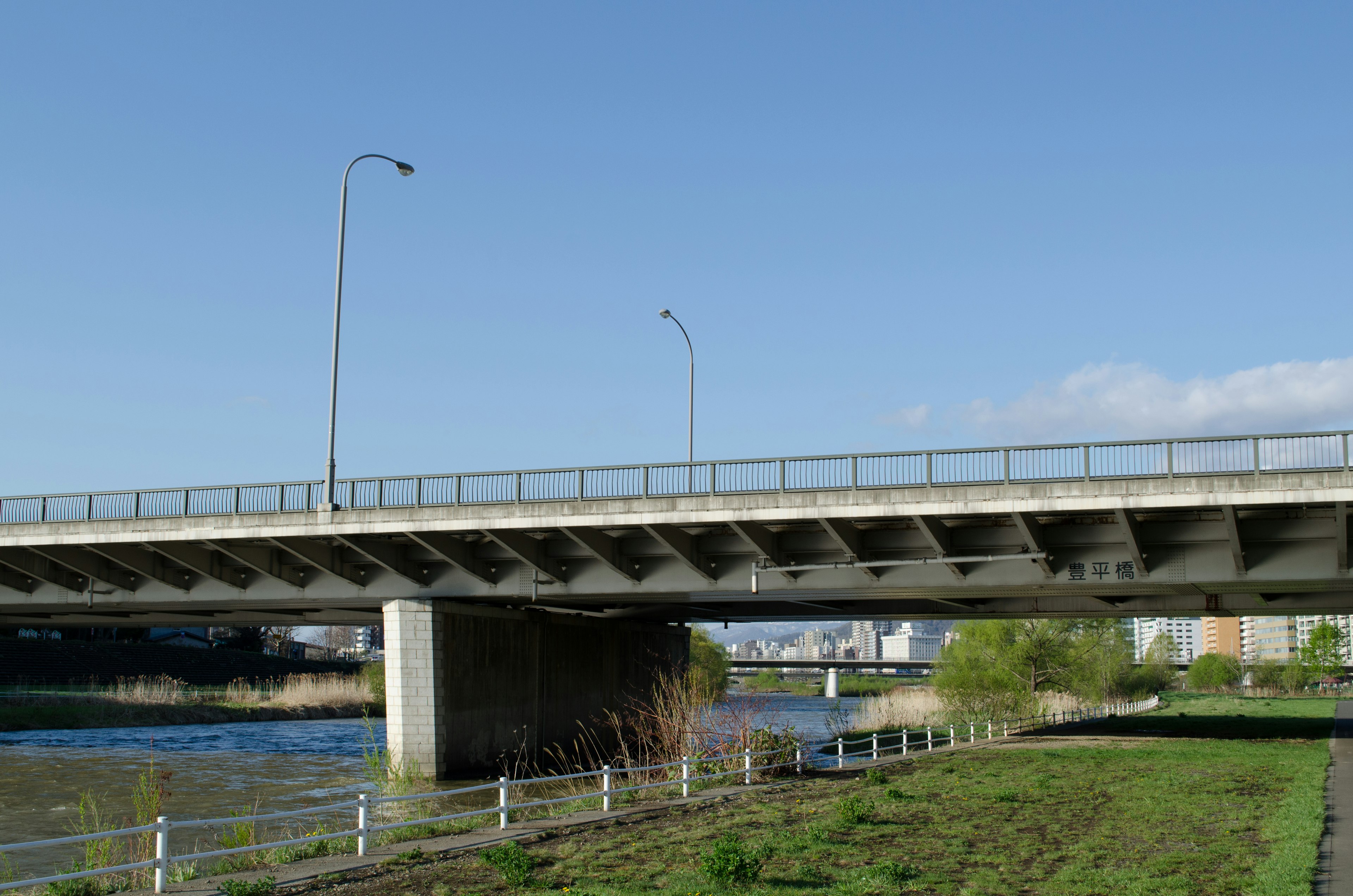 Gambar jembatan beton di bawah langit biru dekat sungai dengan rumput hijau