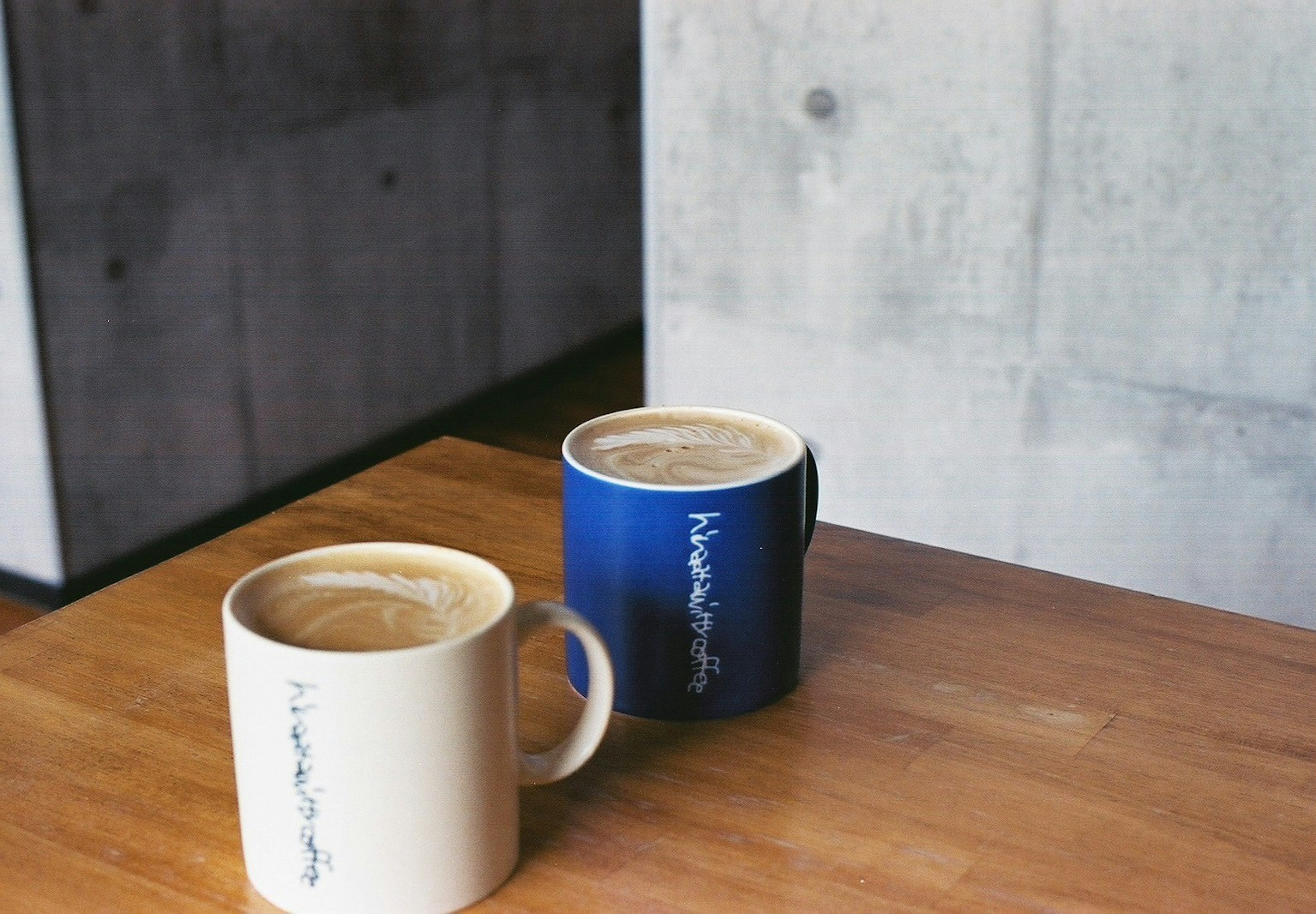 Dua cangkir kopi putih dan biru di atas meja kayu dengan latar belakang dinding beton