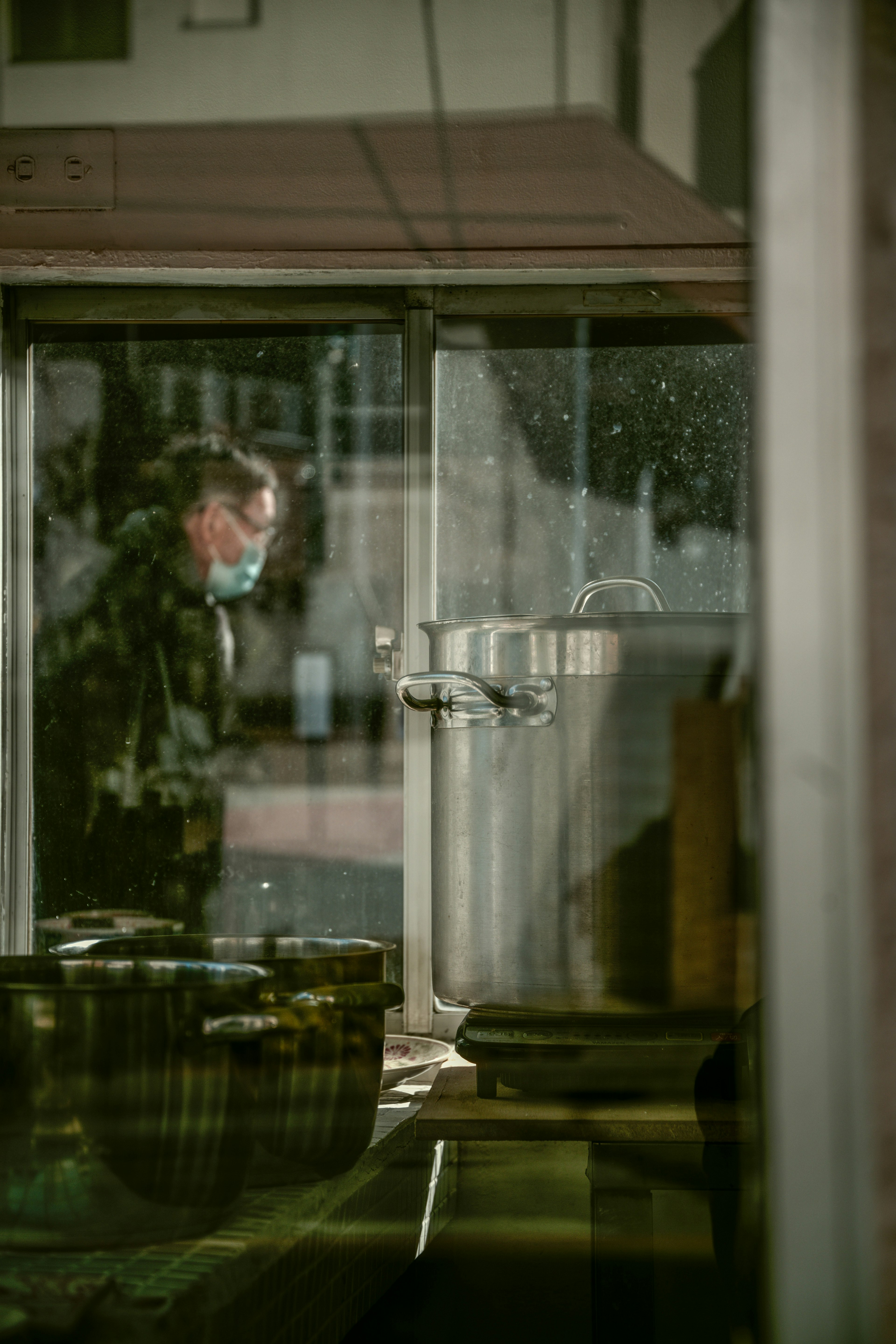 A masked worker seen through a window in a kitchen setting