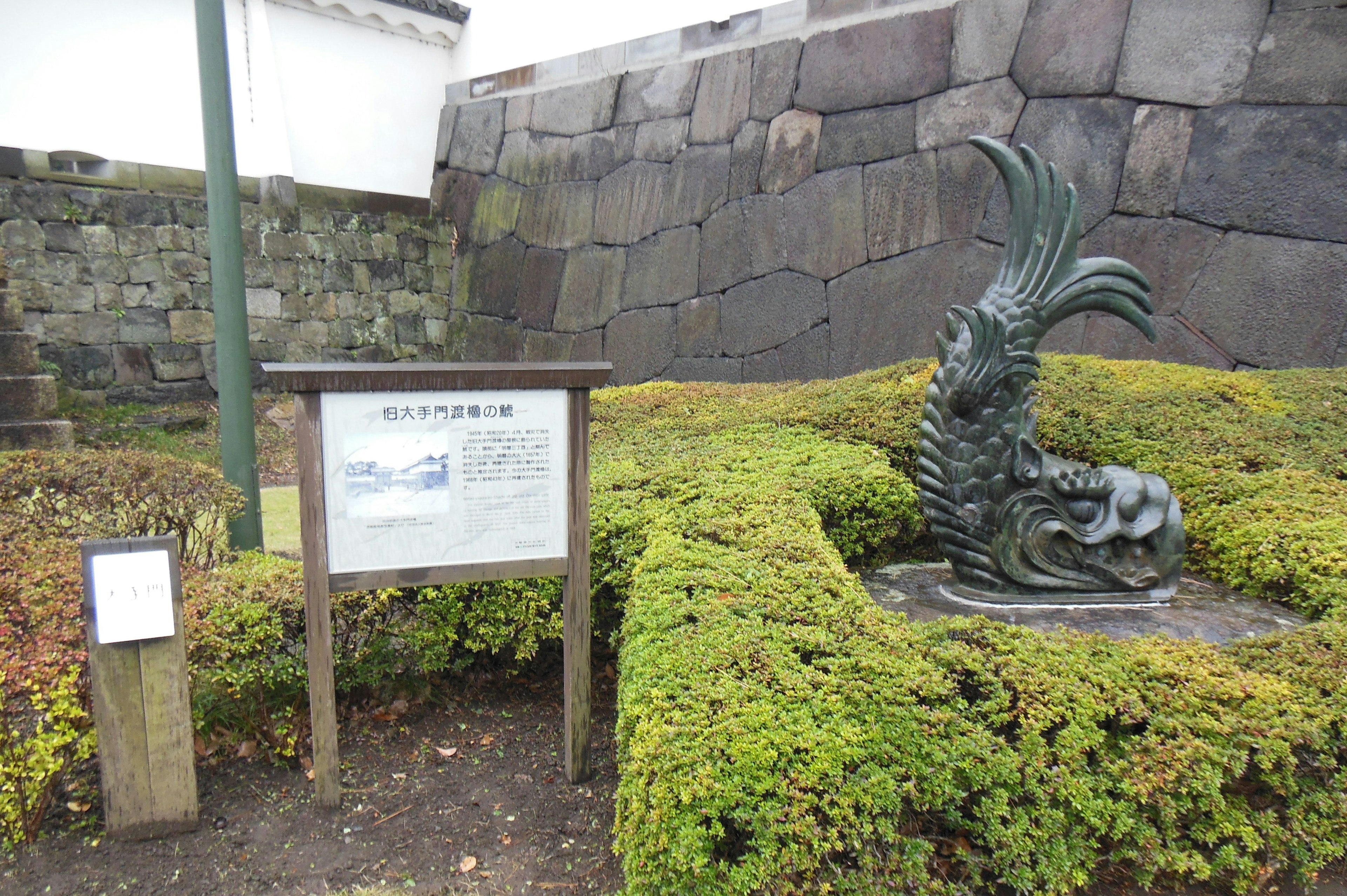 Metallskulptur eines Huhns in einer Parklandschaft mit einem Schild und einer Steinmauer im Hintergrund