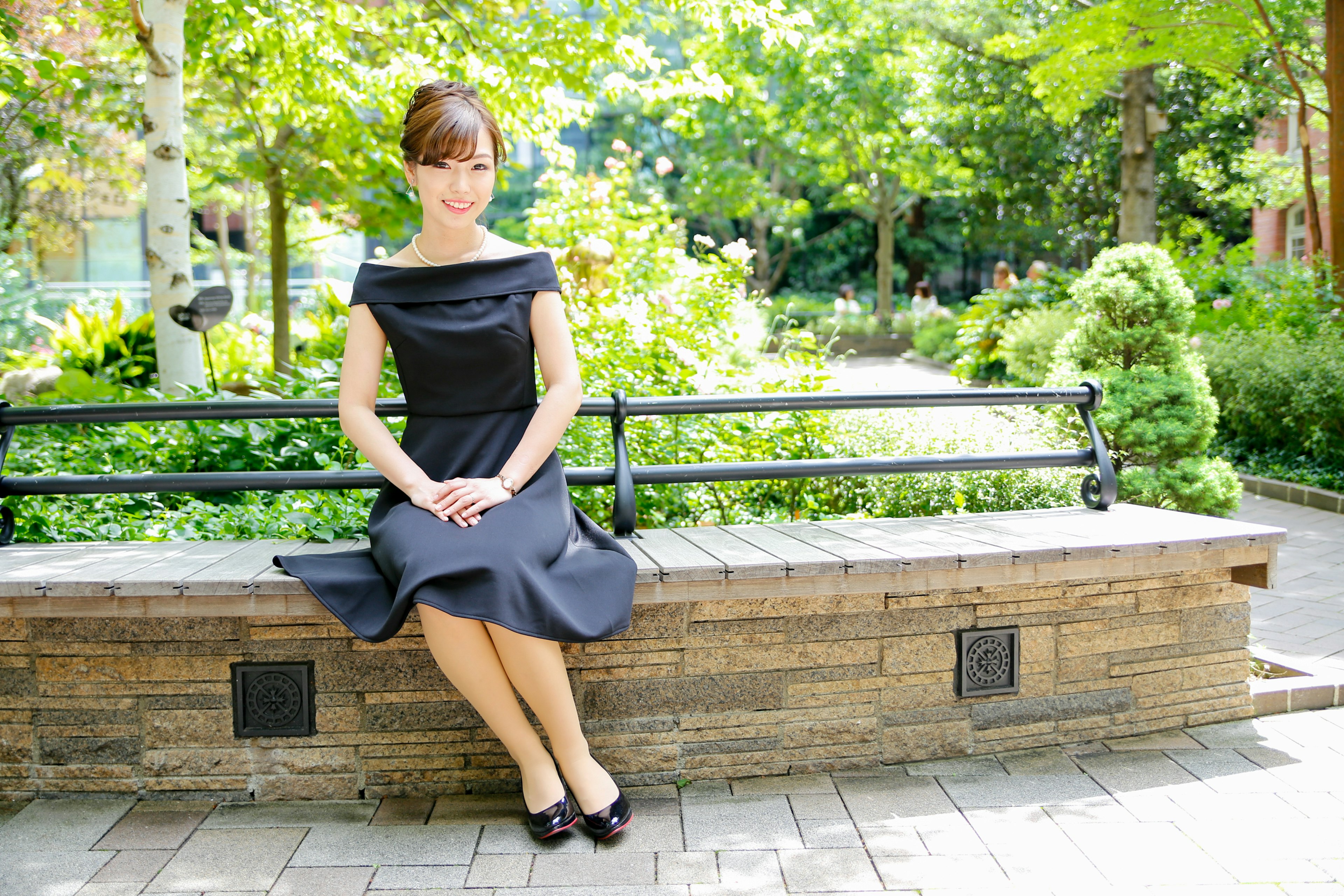 A woman sitting on a park bench wearing a black dress surrounded by lush greenery