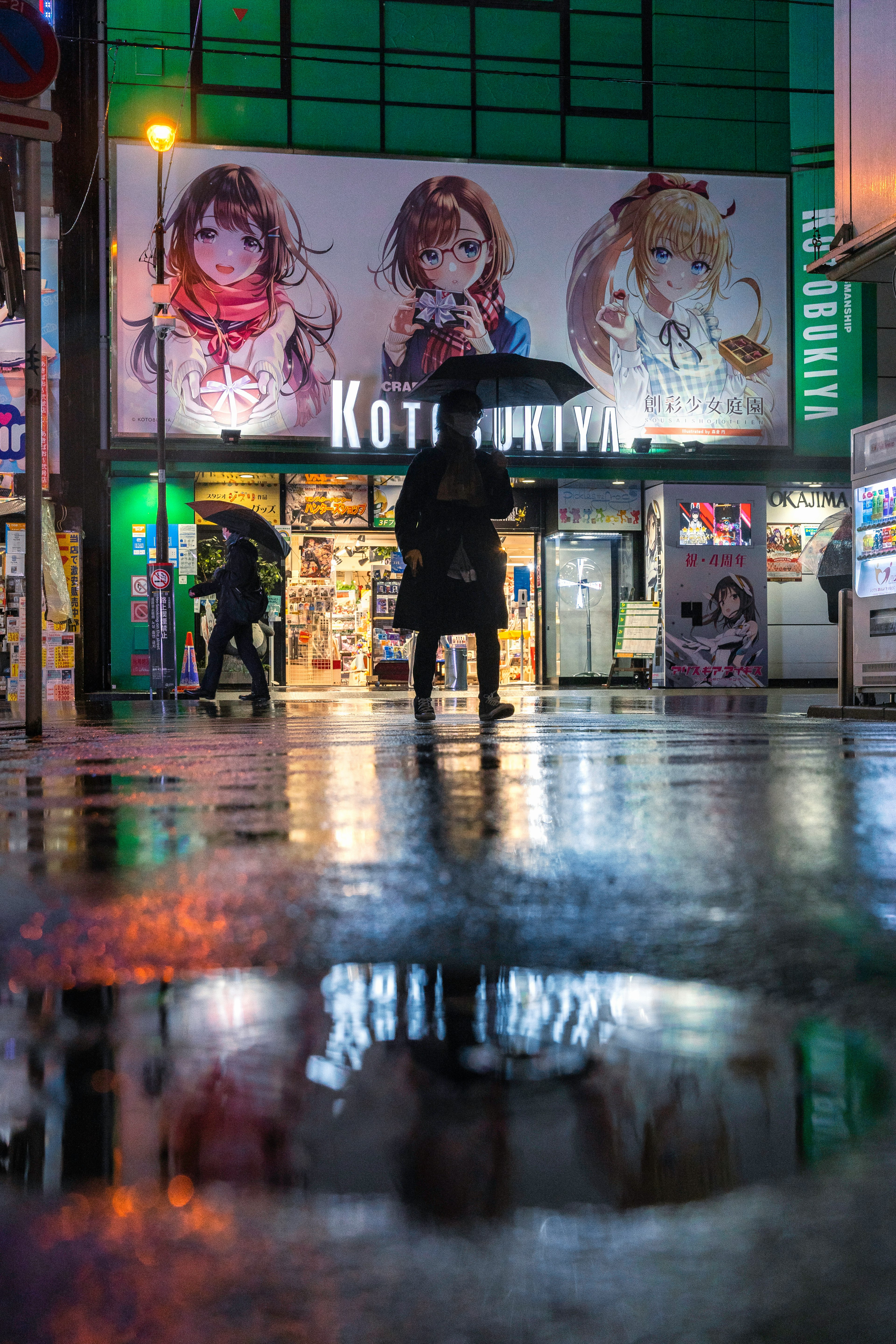 雨の中で傘を持つ人物とアニメ風のポスターがある繁華街の風景