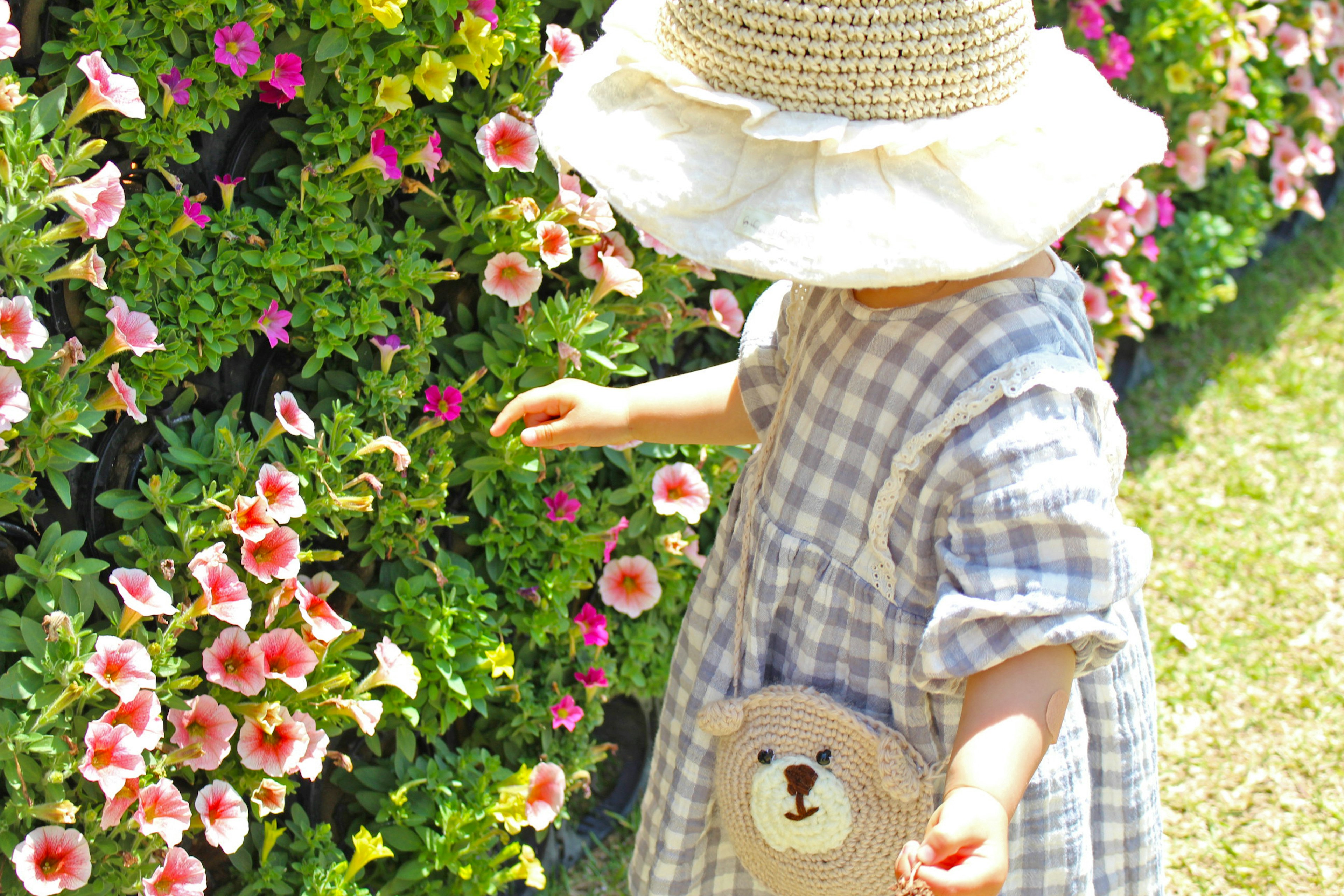 Kind, das vor Blumen spielt, trägt ein kariertes Kleid und einen Sonnenhut