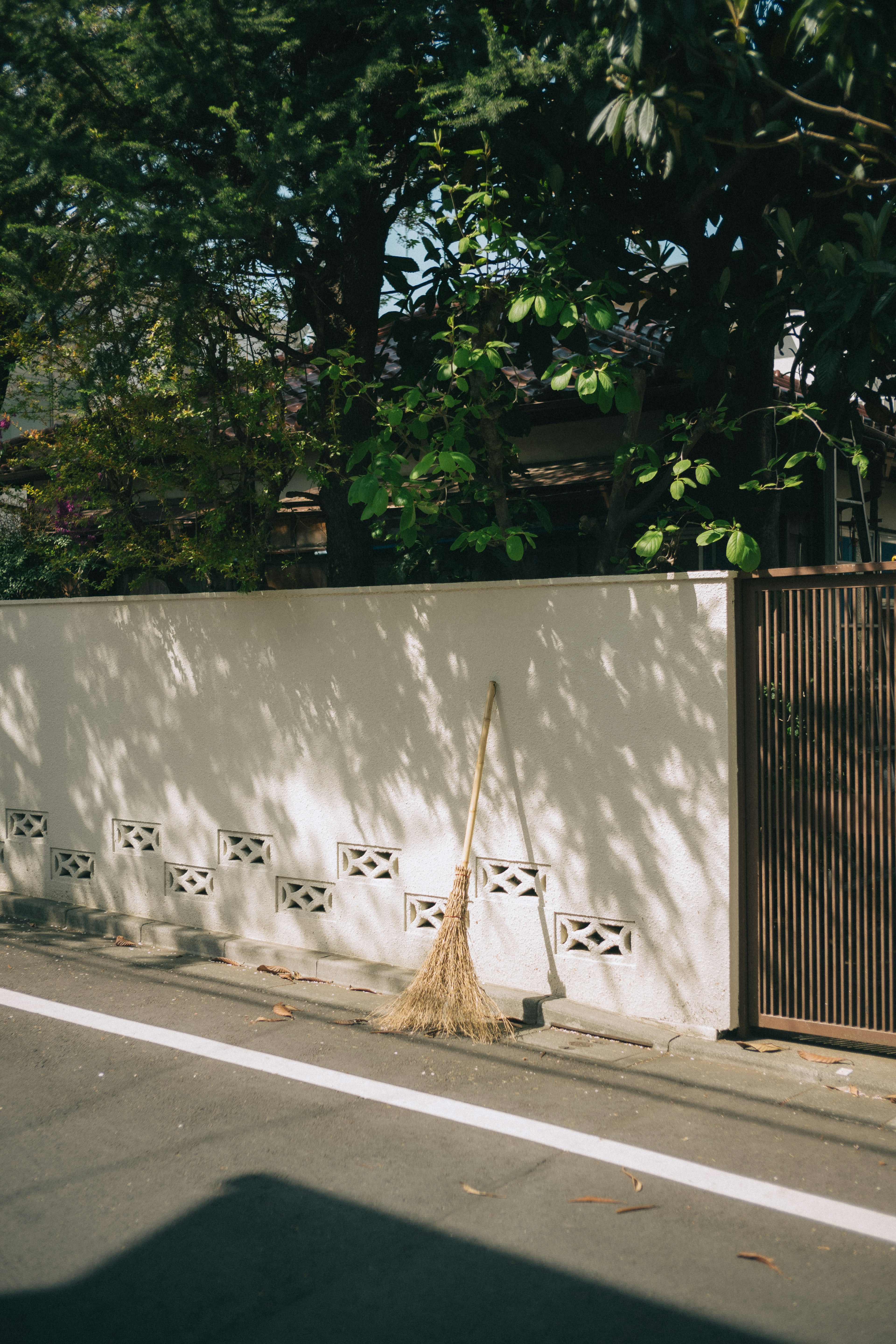 Una escena de calle con una pared blanca y sombras de árboles con una escoba de bambú apoyada