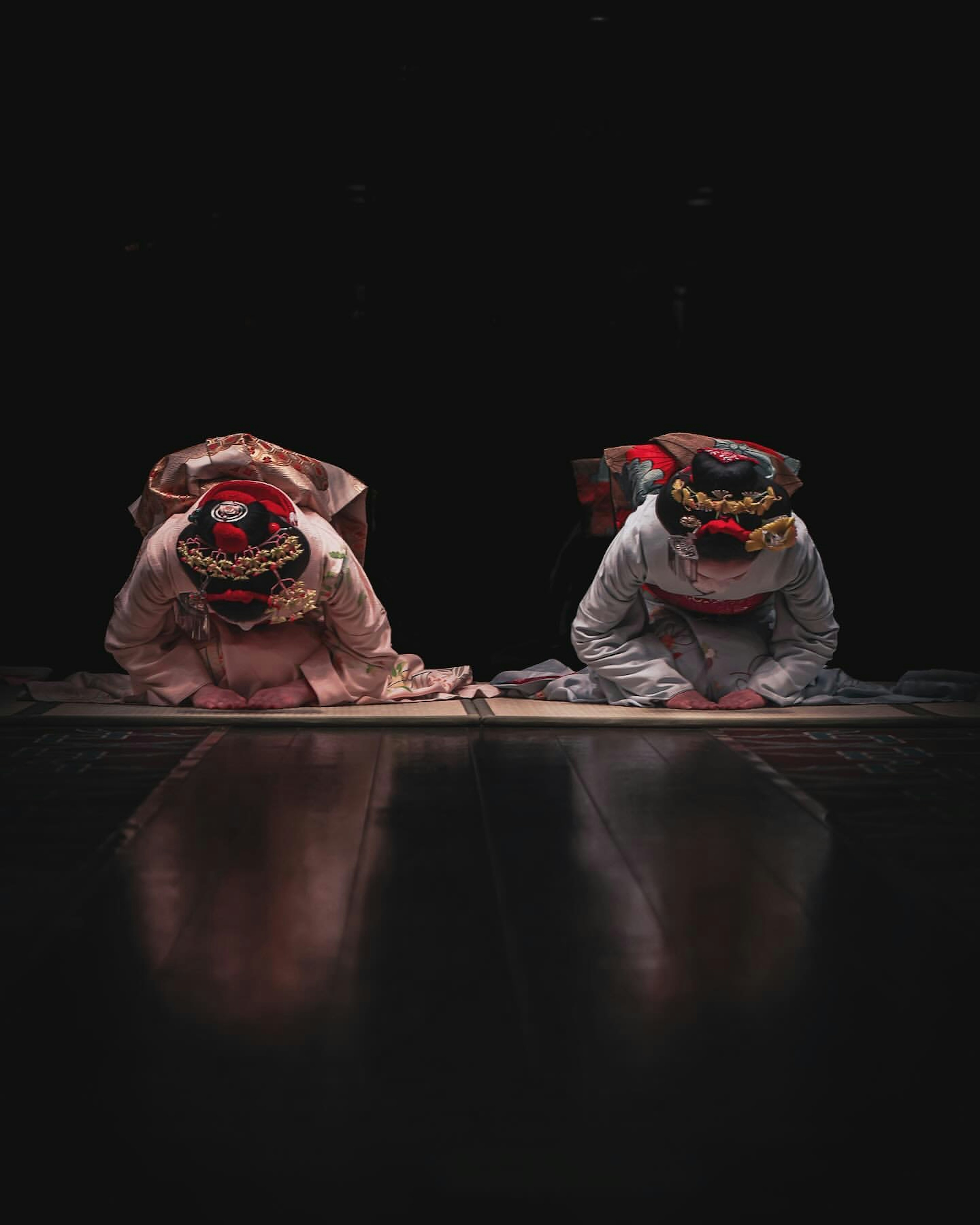 Two performers in traditional costumes kneeling and bowing in front of a dark background