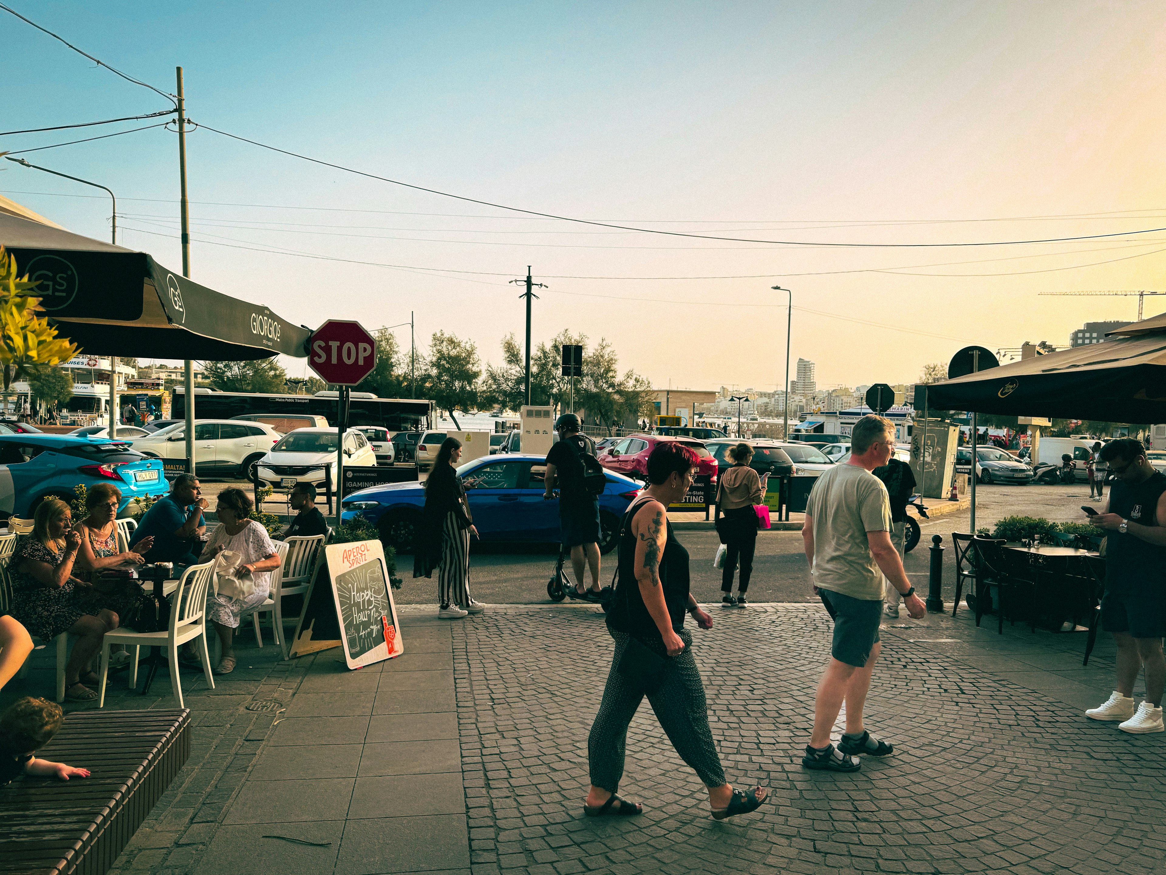 Scena vivace di un caffè all'aperto con persone che si radunano al tramonto e auto sullo sfondo