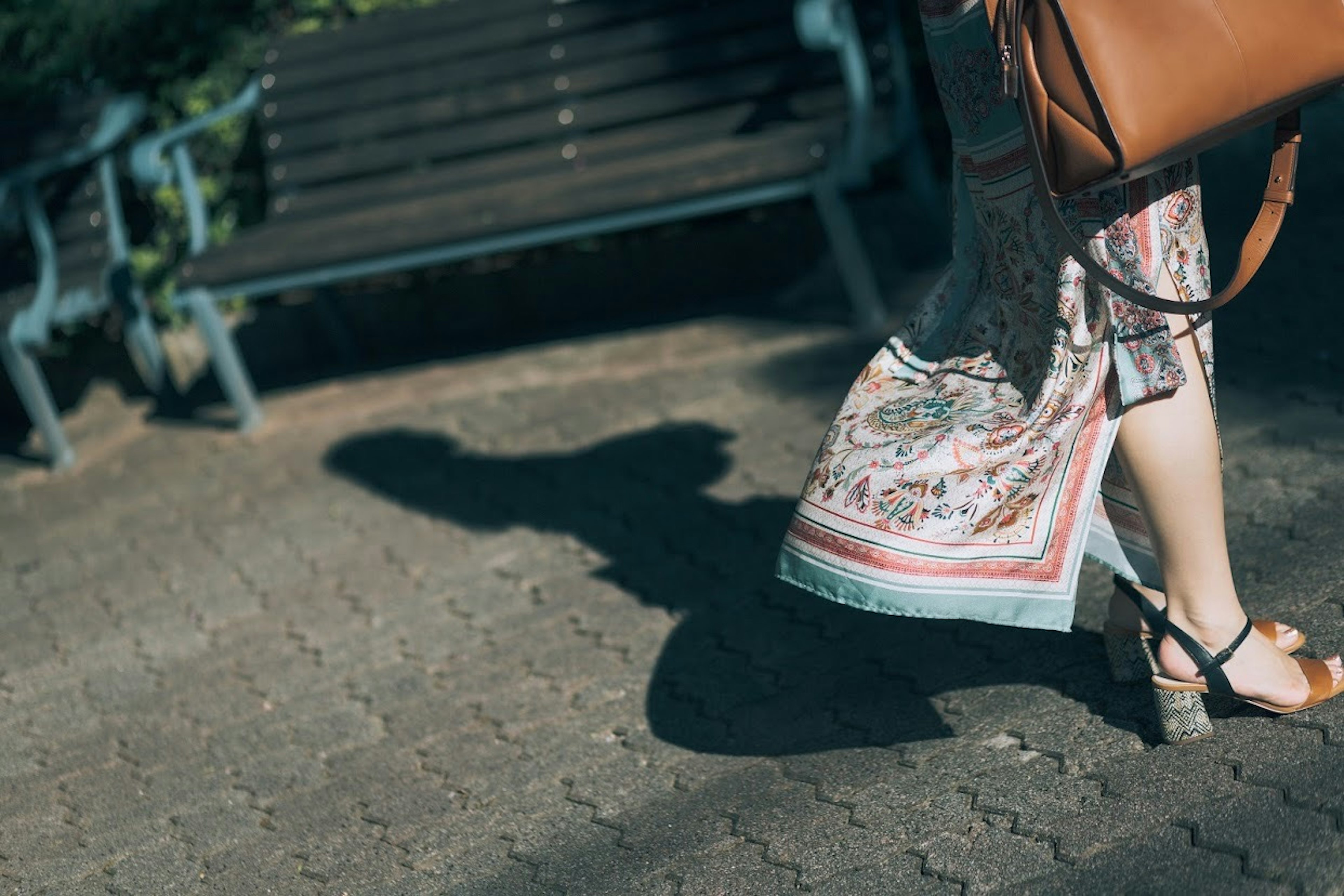 Una mujer caminando en sandalias con un vestido estampado y un bolso proyectando una sombra