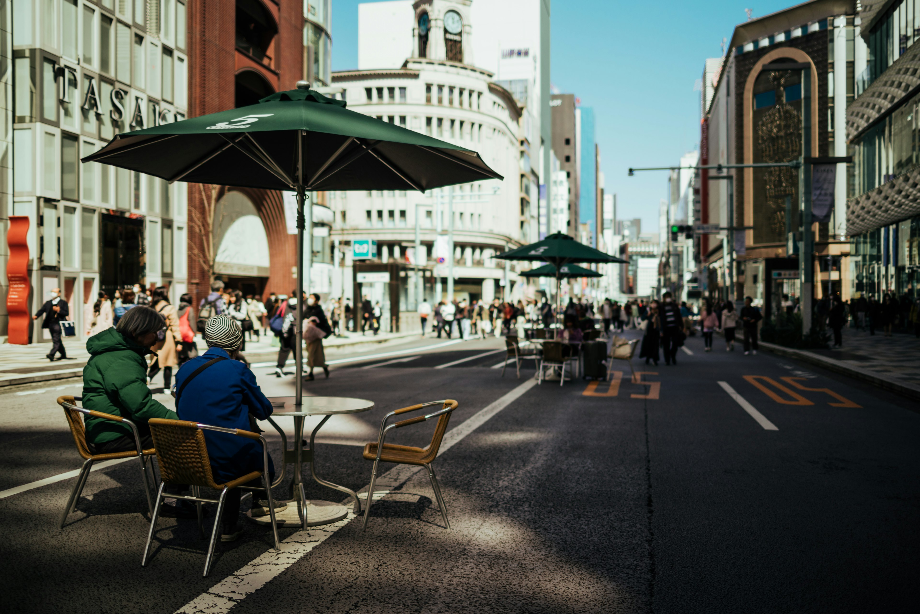 Two people sitting at a table with umbrellas on a busy street filled with pedestrians