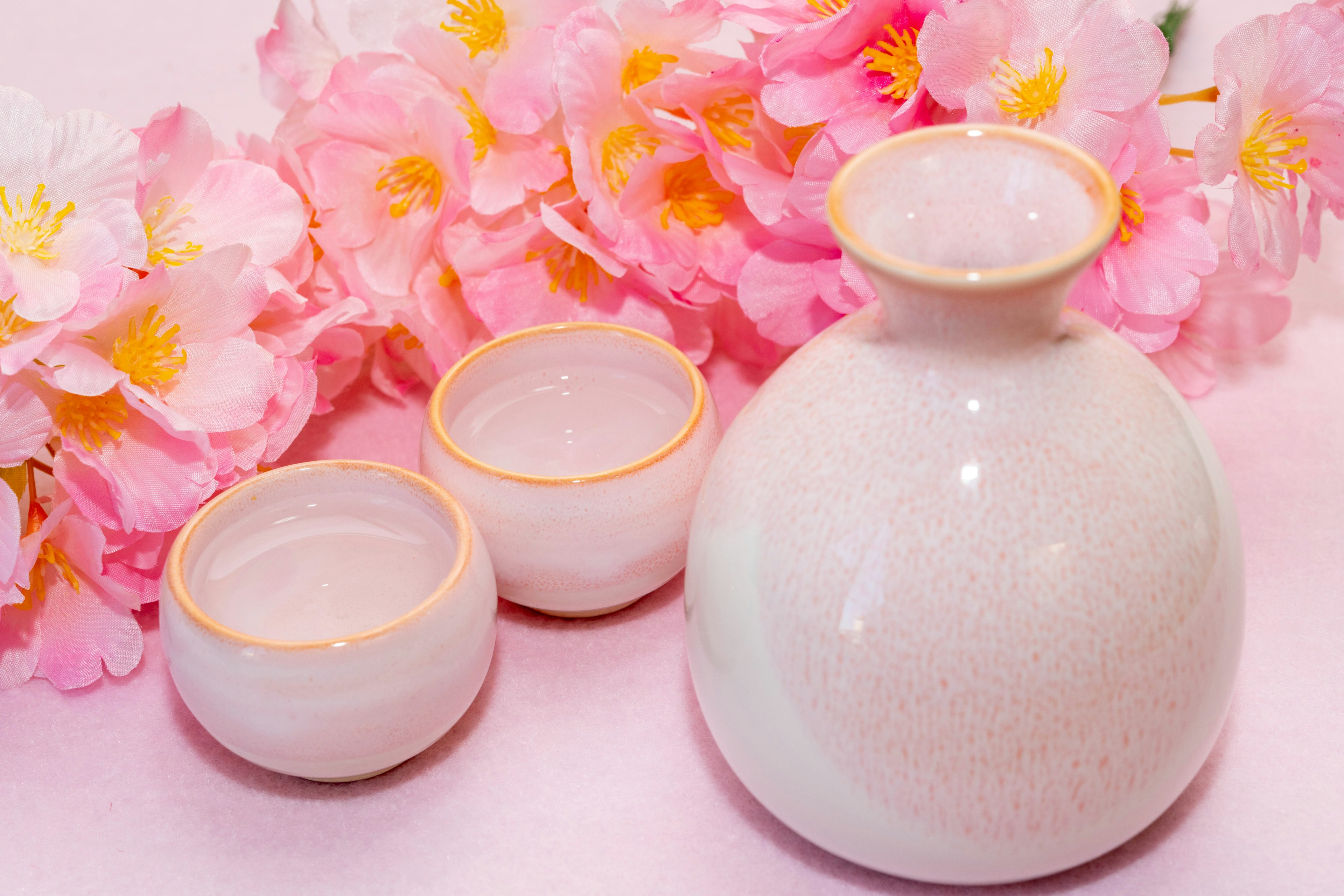 Un set di sakè in ceramica bianca con due piccole tazze accanto a uno sfondo di fiori di ciliegio rosa