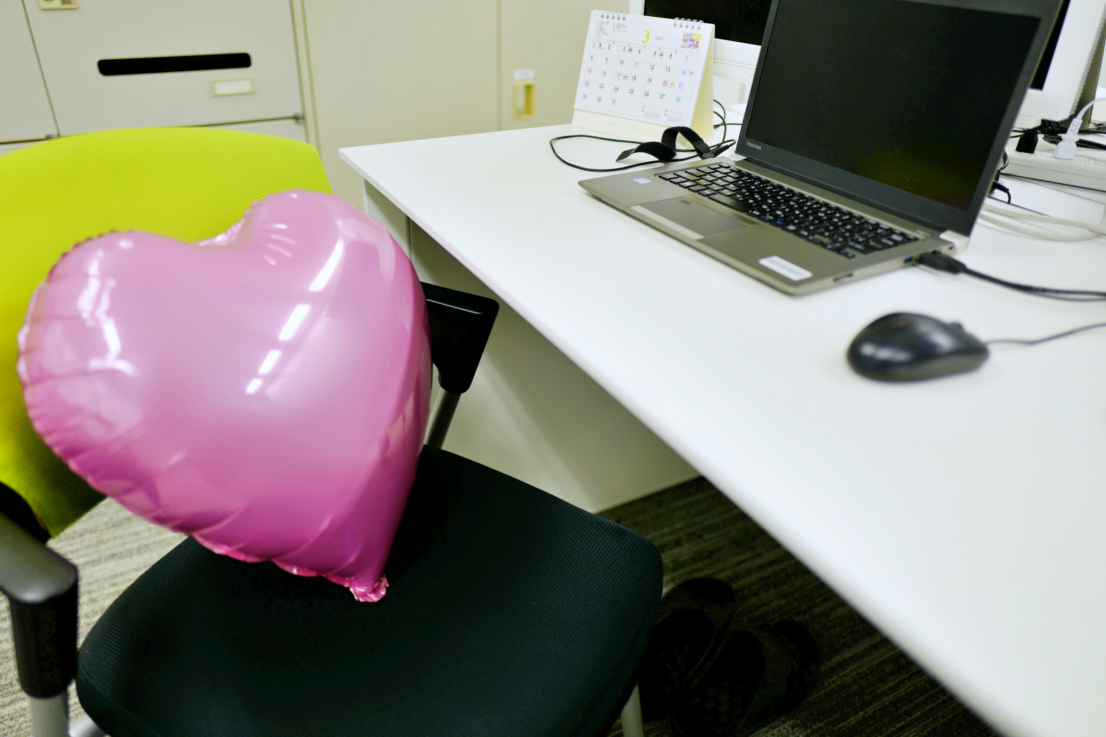 Pink heart-shaped balloon resting on a green chair next to a desk with a computer and mouse