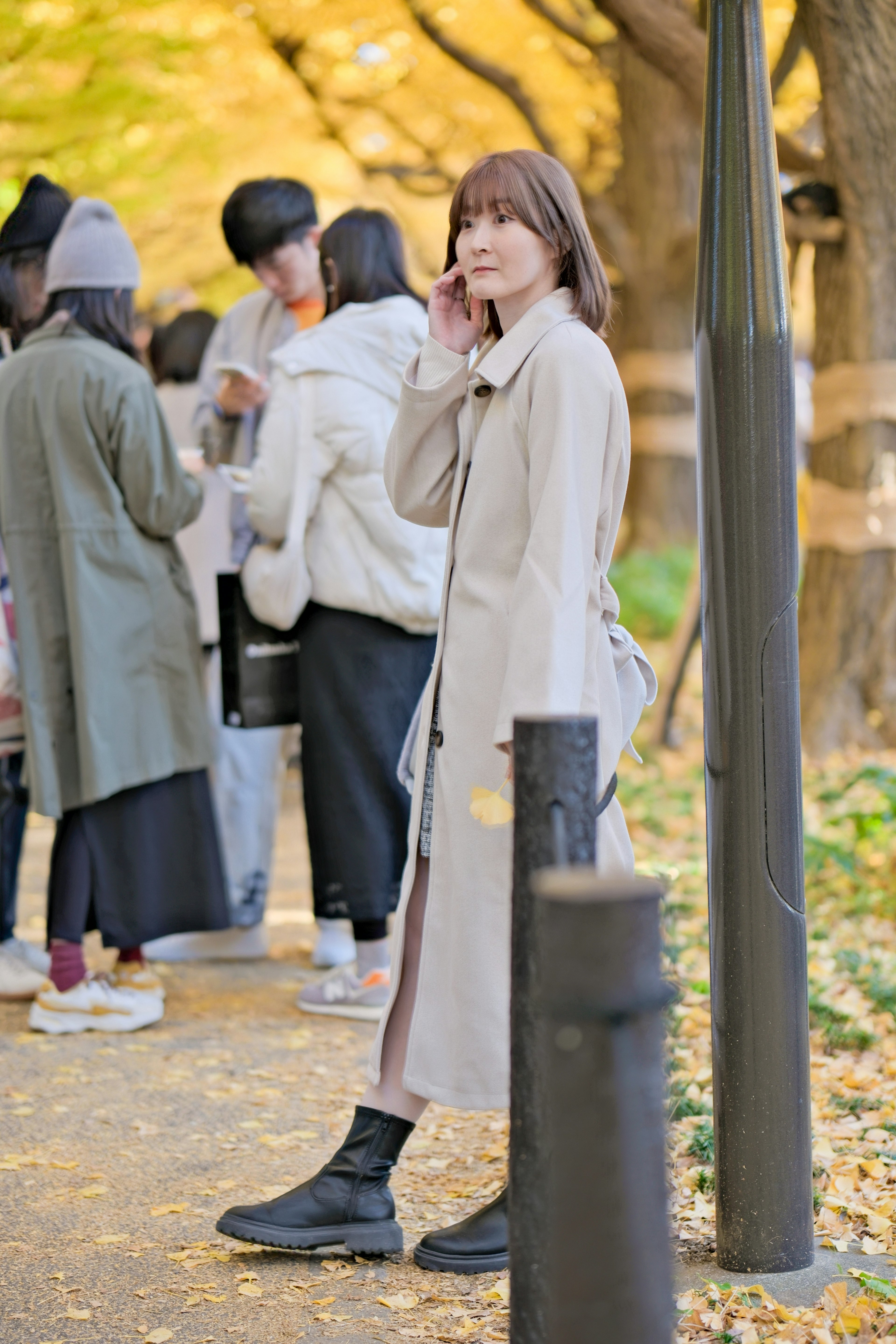 Una mujer hablando por teléfono en un camino del parque rodeada de personas con hojas de otoño esparcidas