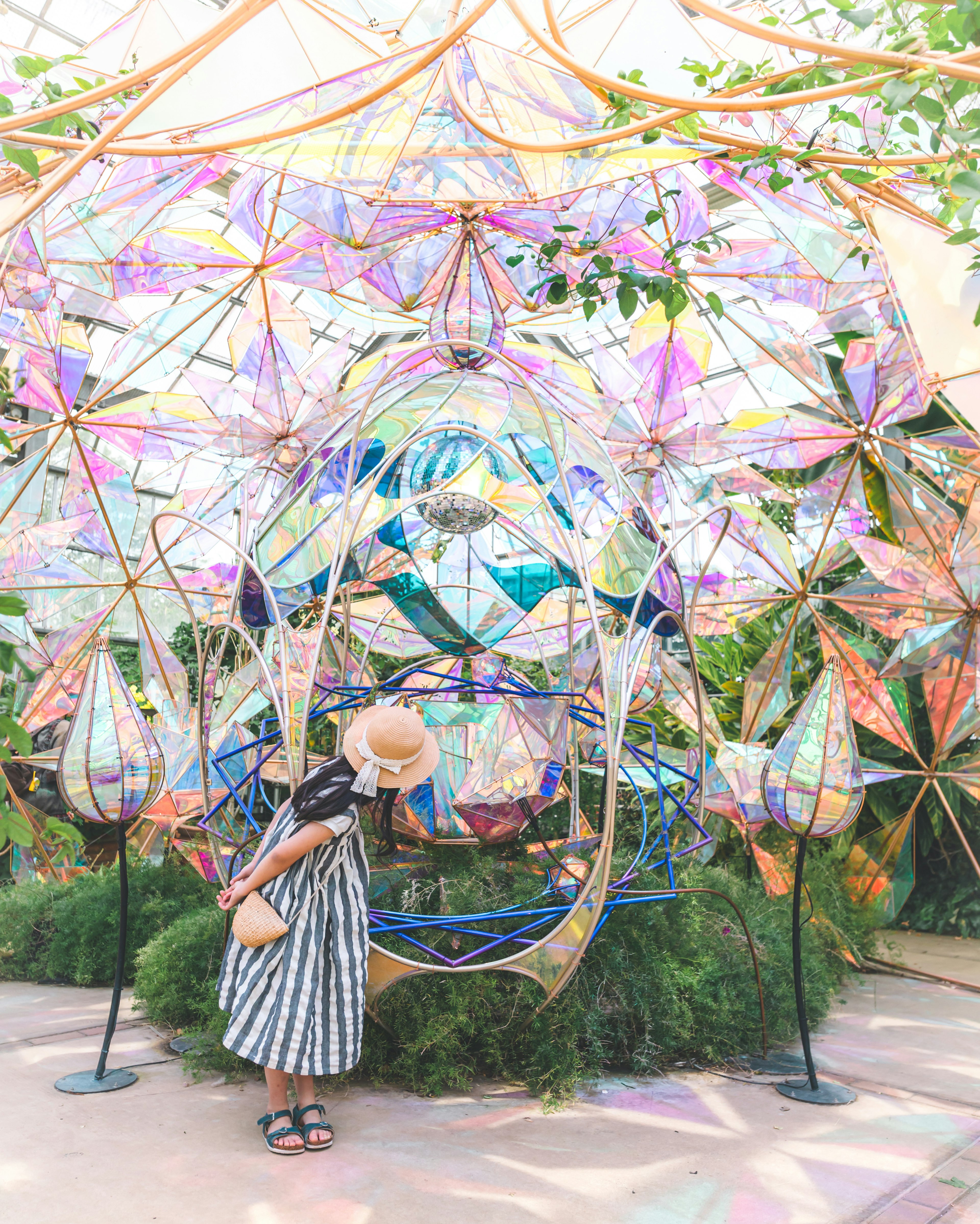 Una mujer con un vestido a rayas y sombrero está de pie frente a una colorida instalación artística con diseños intrincados