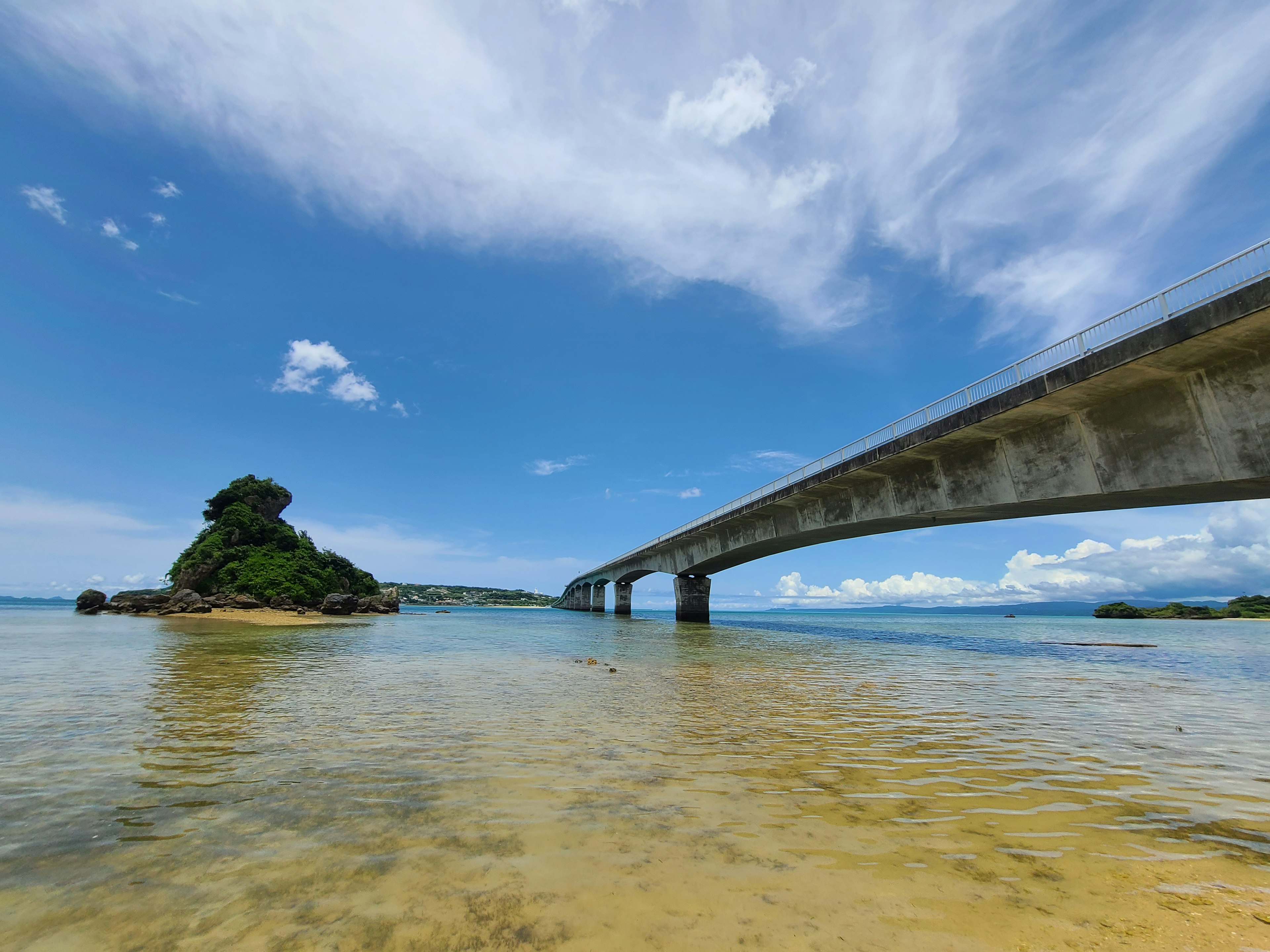 Pemandangan jembatan yang membentang di atas air jernih menuju sebuah pulau di bawah langit biru