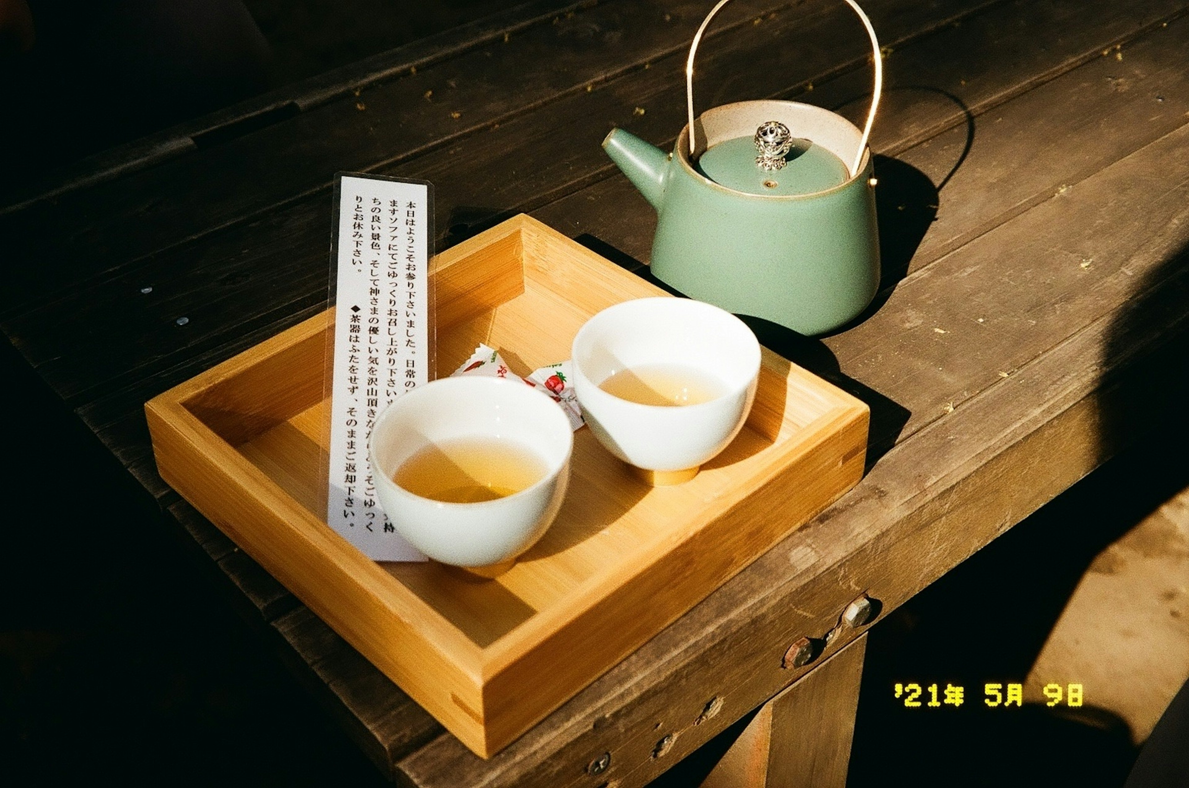 A wooden tray with two white teacups and a green teapot with a note beside them