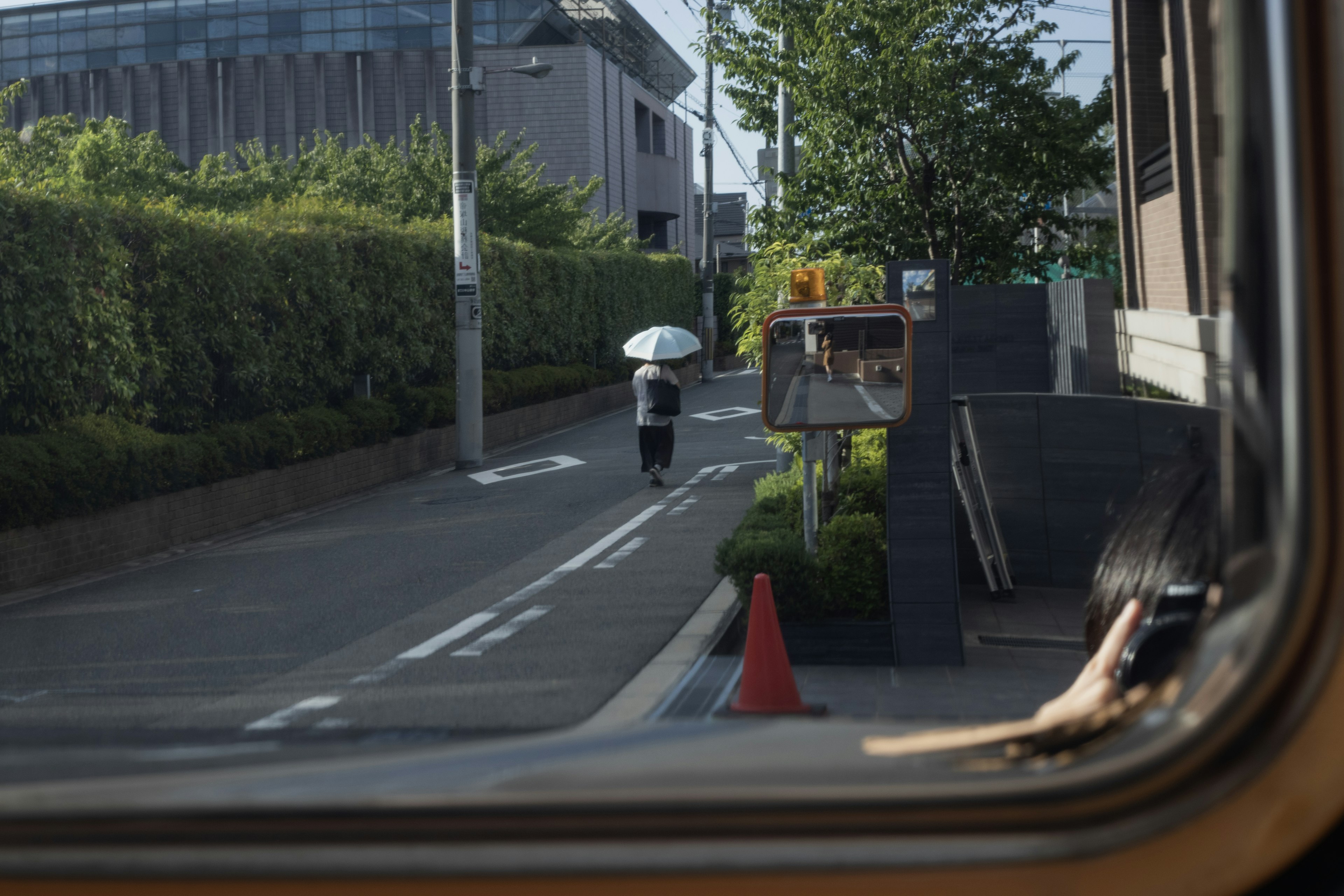 窓越しに見える通りを歩く人傘をさしている緑の木々と建物が背景