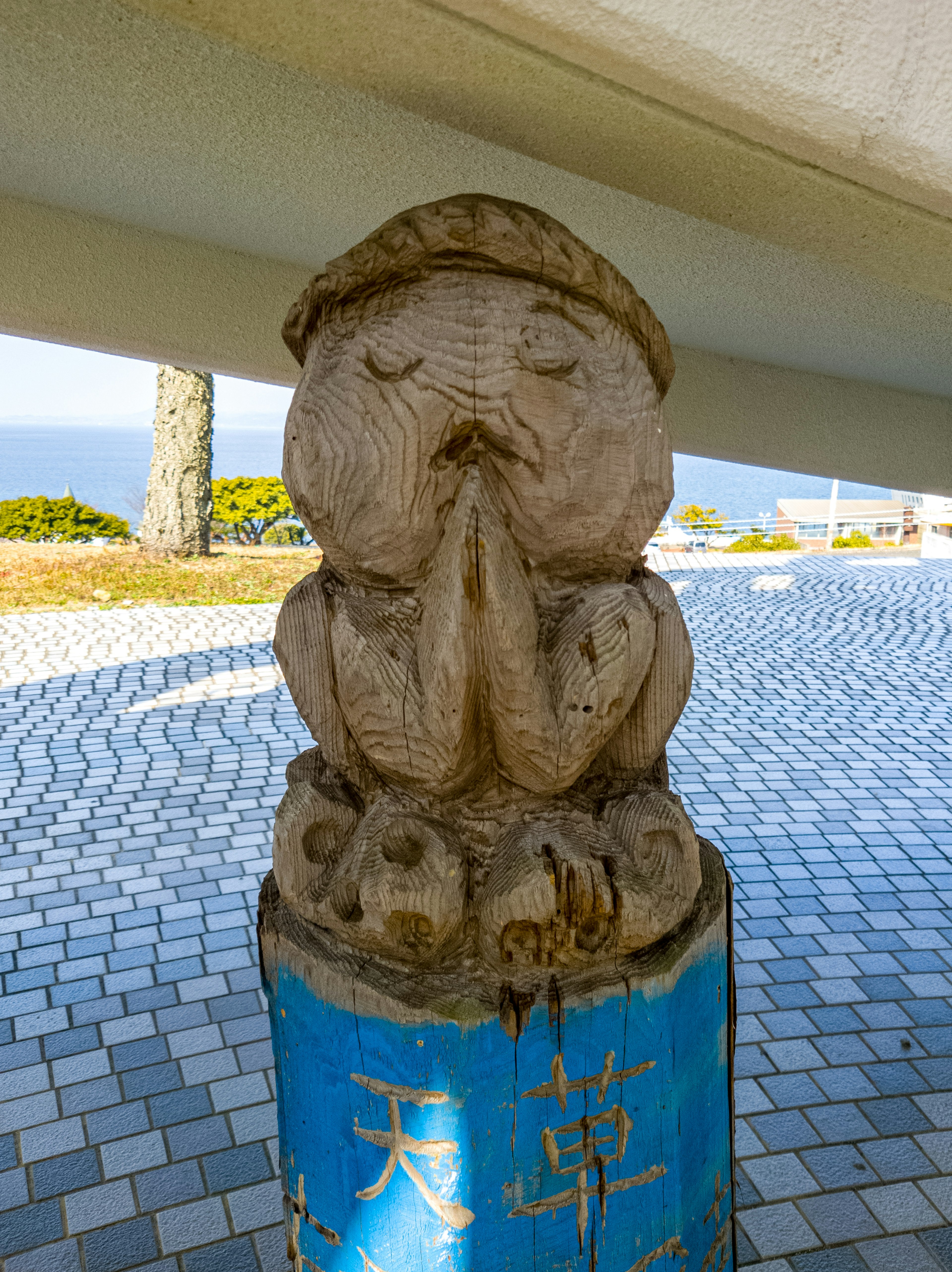 Statua in legno di un personaggio in preghiera con una base blu che mostra caratteri kanji