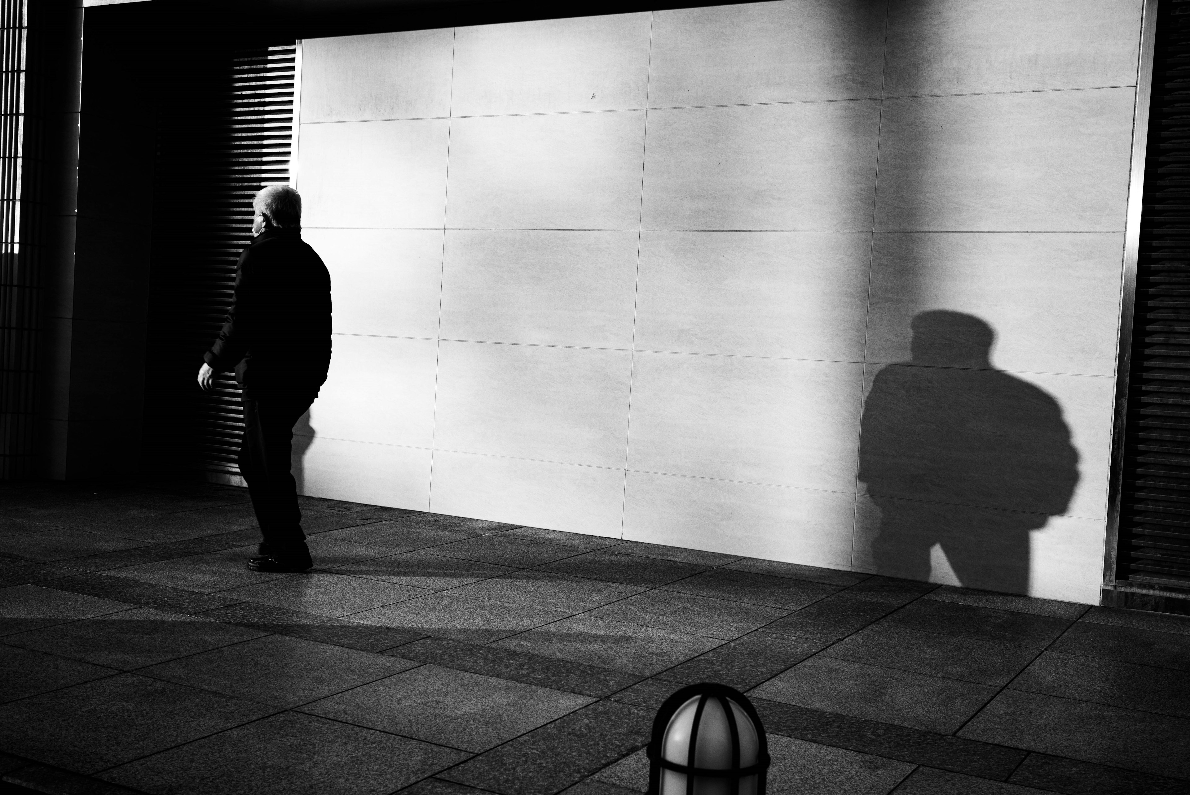 Una escena callejera en blanco y negro de alto contraste con una persona caminando y su sombra proyectada en una pared