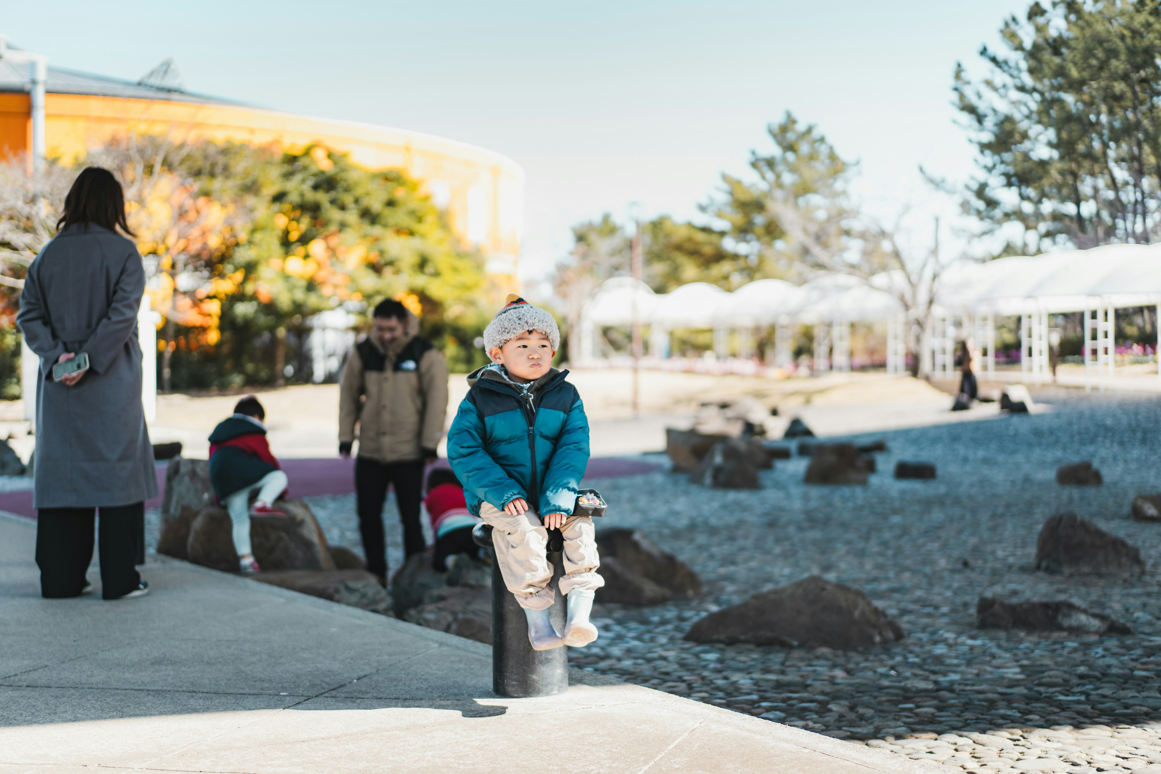 A child in a blue jacket sitting on a post in a park with other children playing