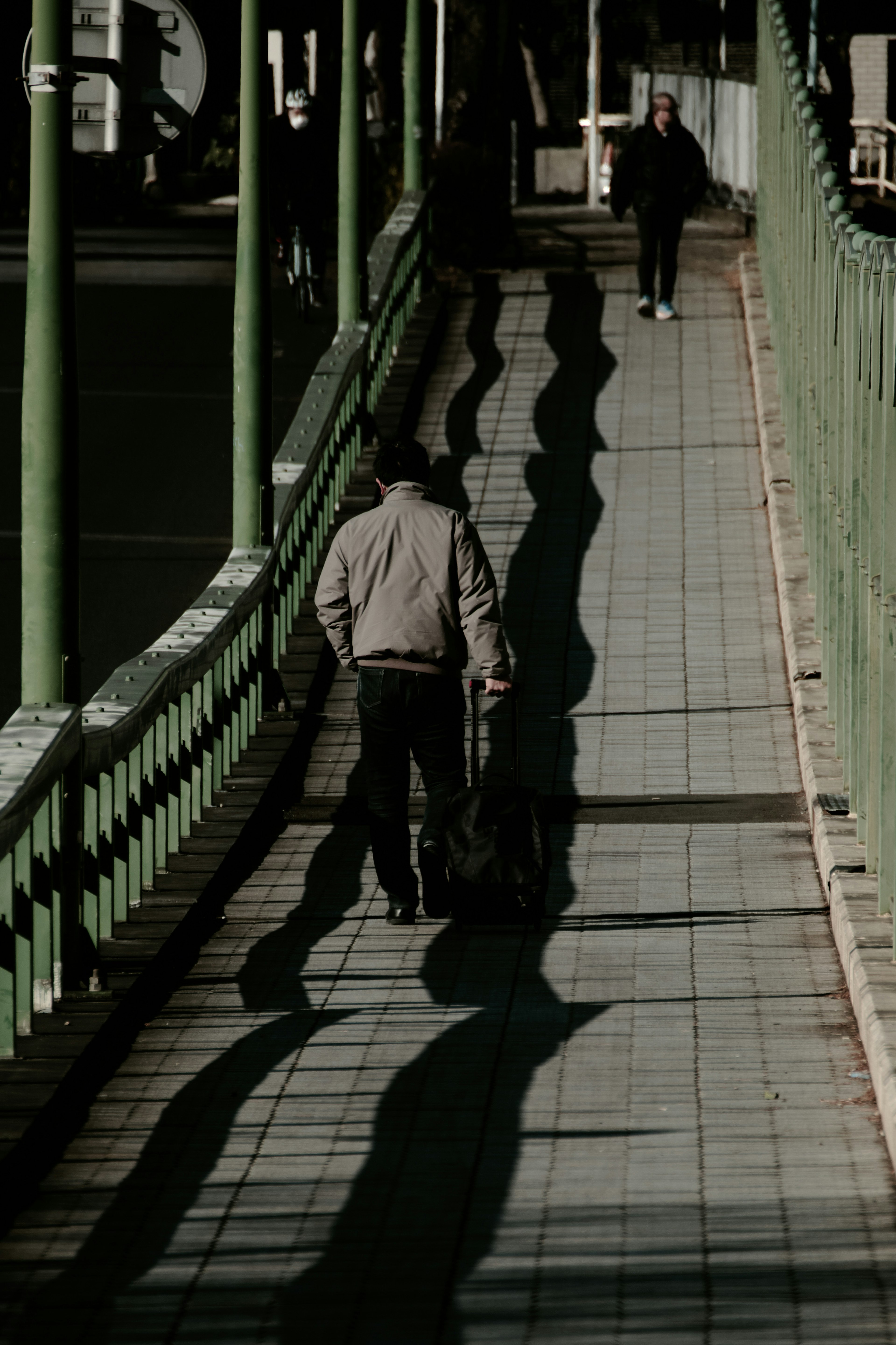 Una persona che cammina lungo un marciapiede con recinzione verde e lunghe ombre