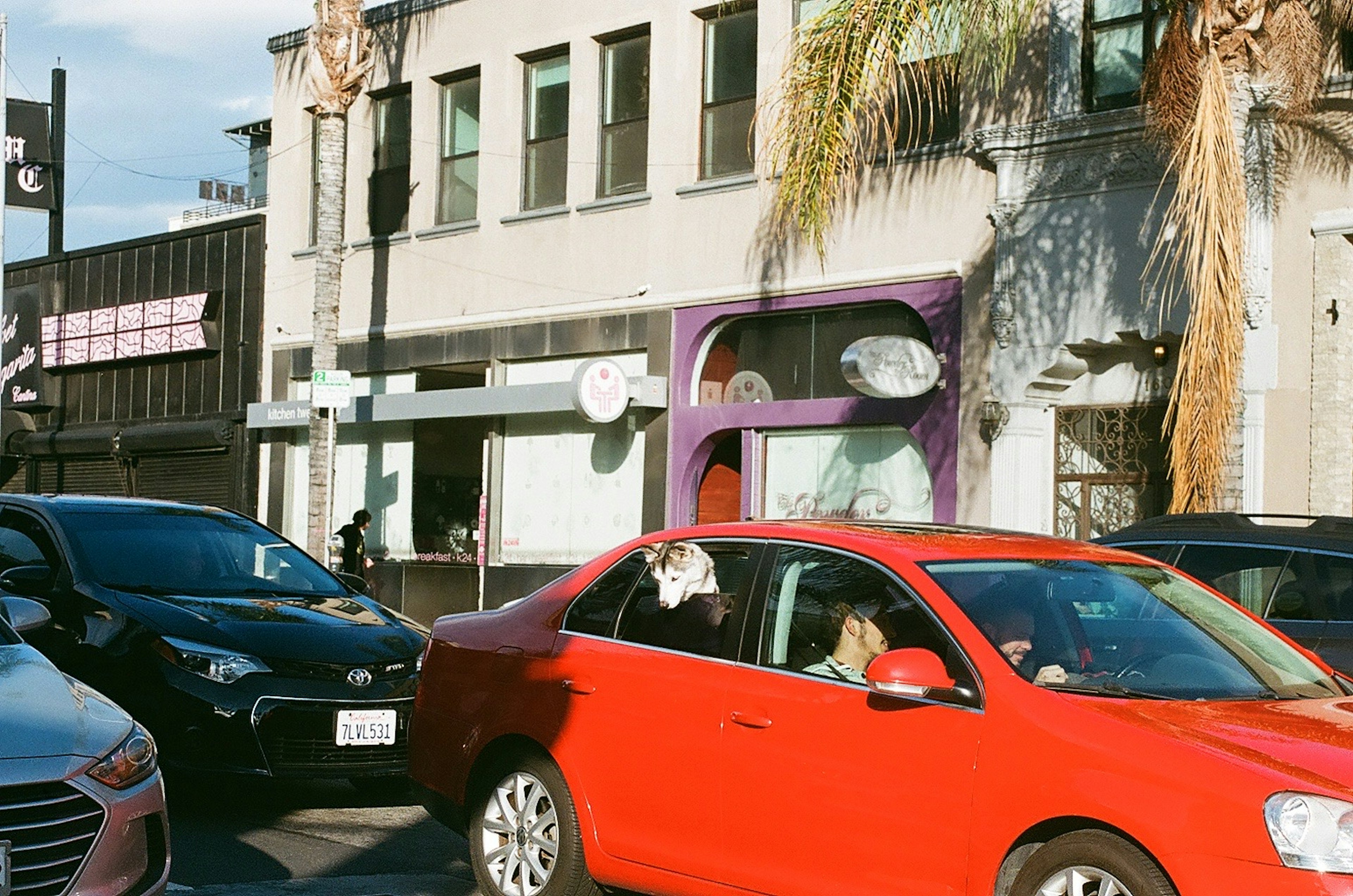 A red car in traffic with a palm tree and a purple storefront