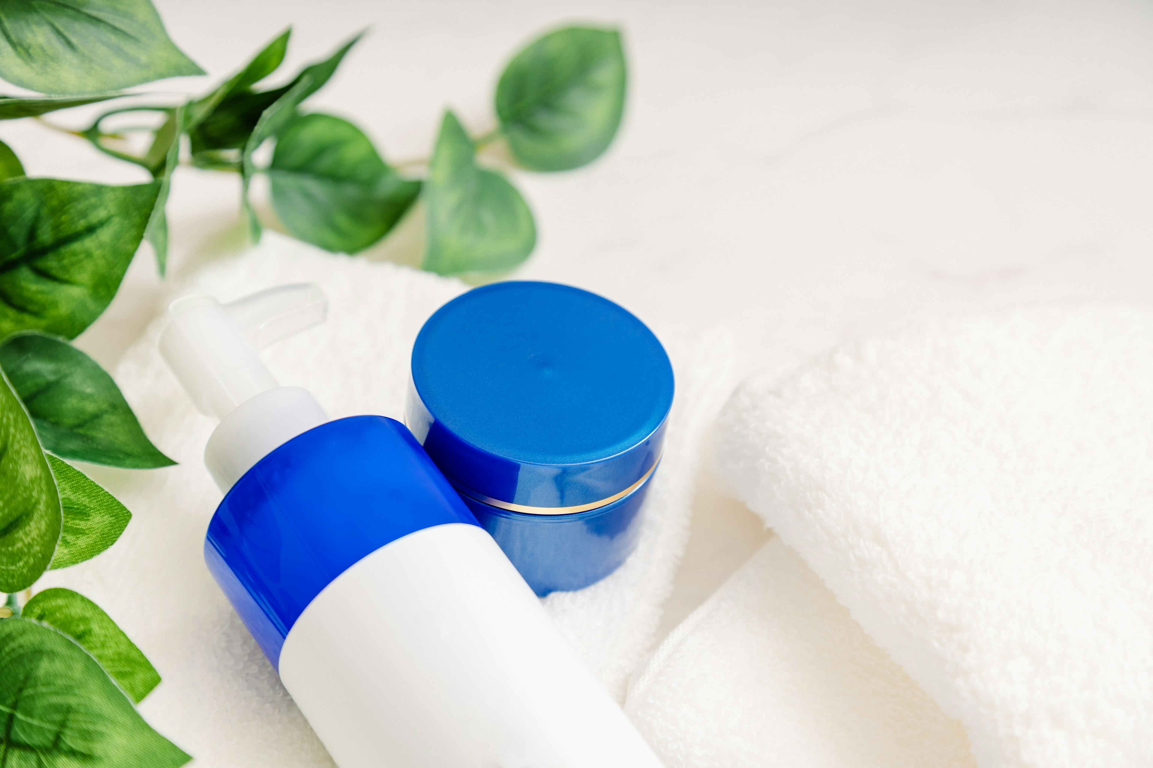 A blue capped cream jar and a spray bottle placed on a towel with green leaves in the background