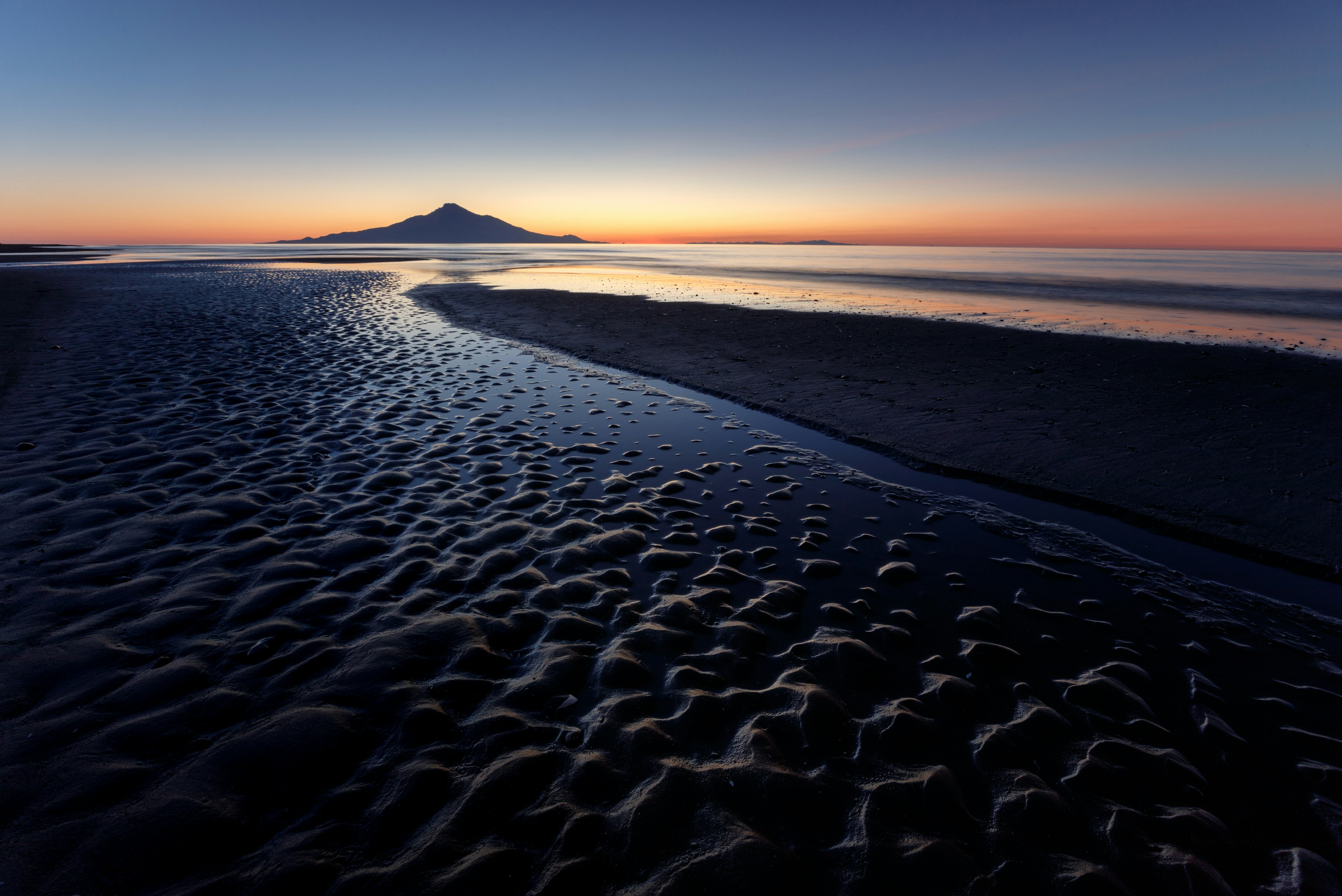 A tranquil seascape at sunset featuring rippled sand and a distant mountain