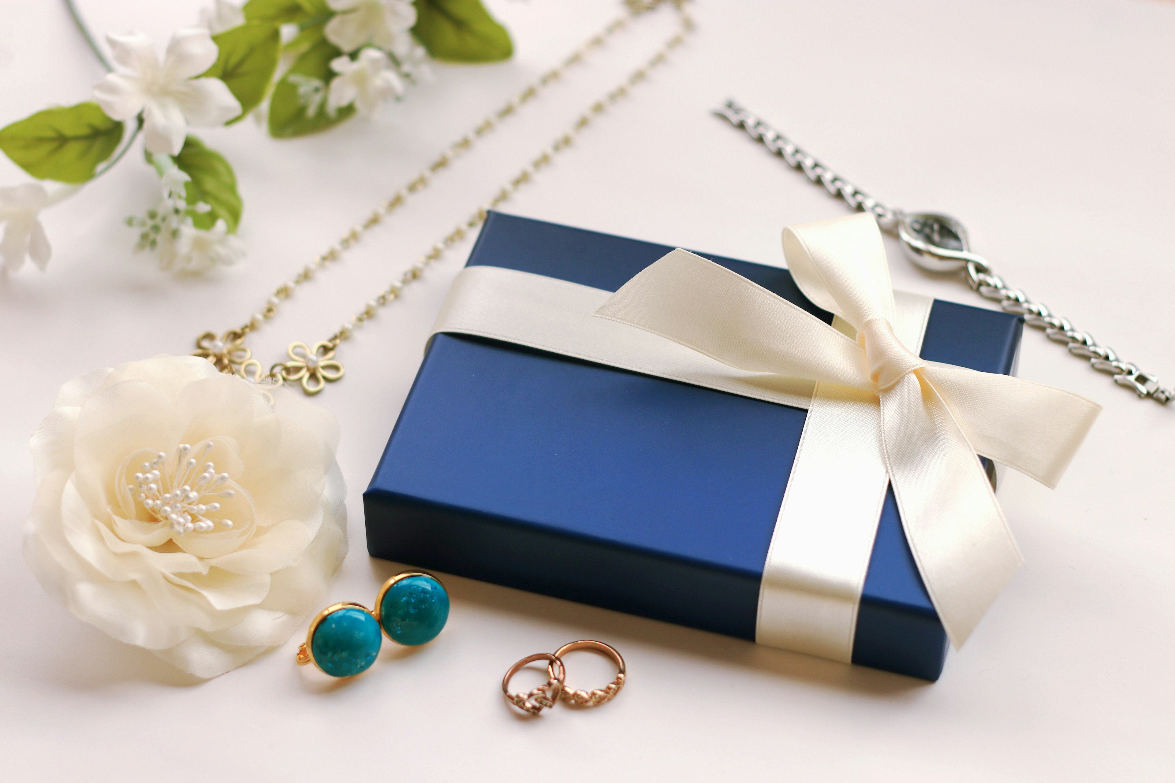 Boîte cadeau bleue avec ruban blanc entourée de bijoux incluant un collier, des boucles d'oreilles et une fleur