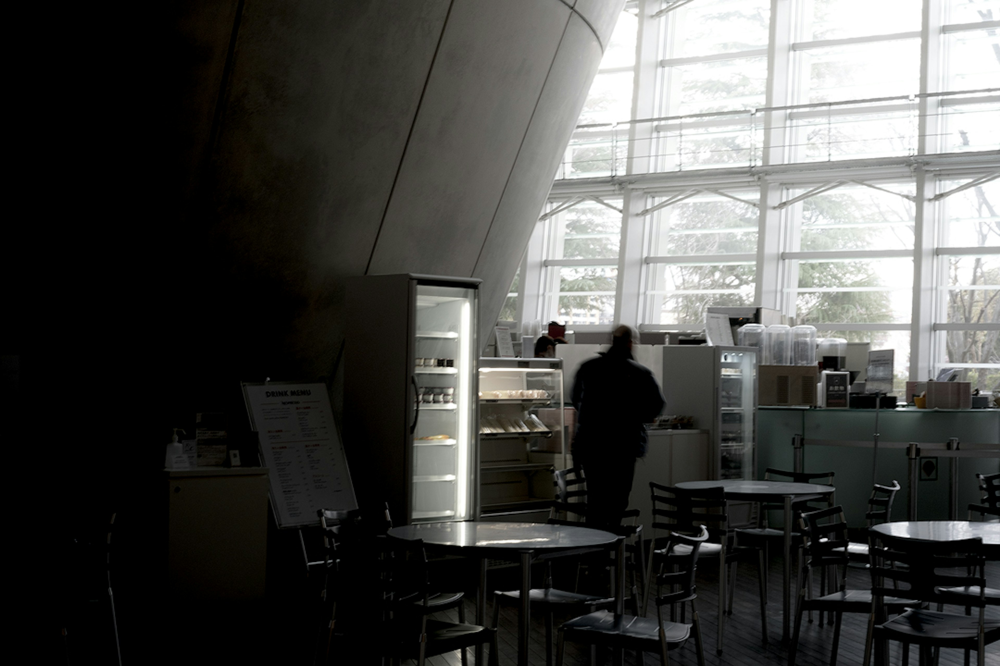 Interior of a cafe with large bright windows featuring a person in shadow near a refrigerator