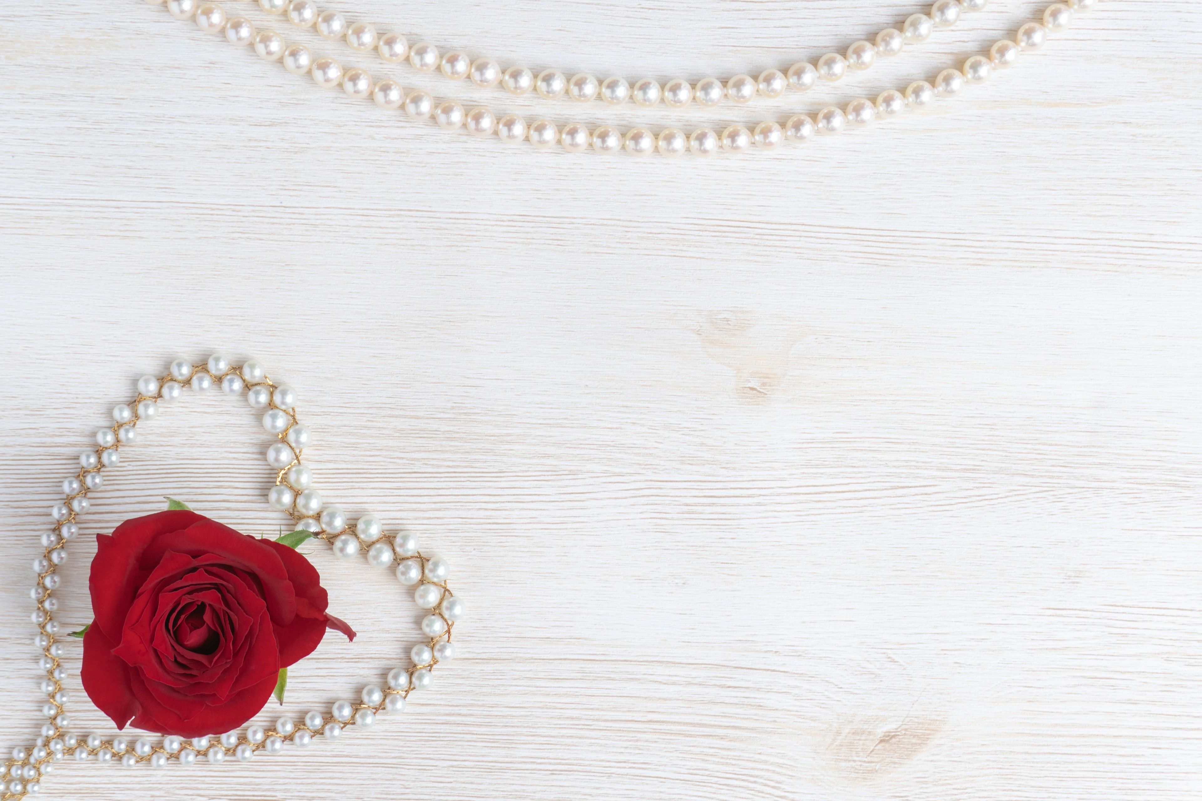 Una rosa rossa e una collana di perle disposte su uno sfondo in legno bianco