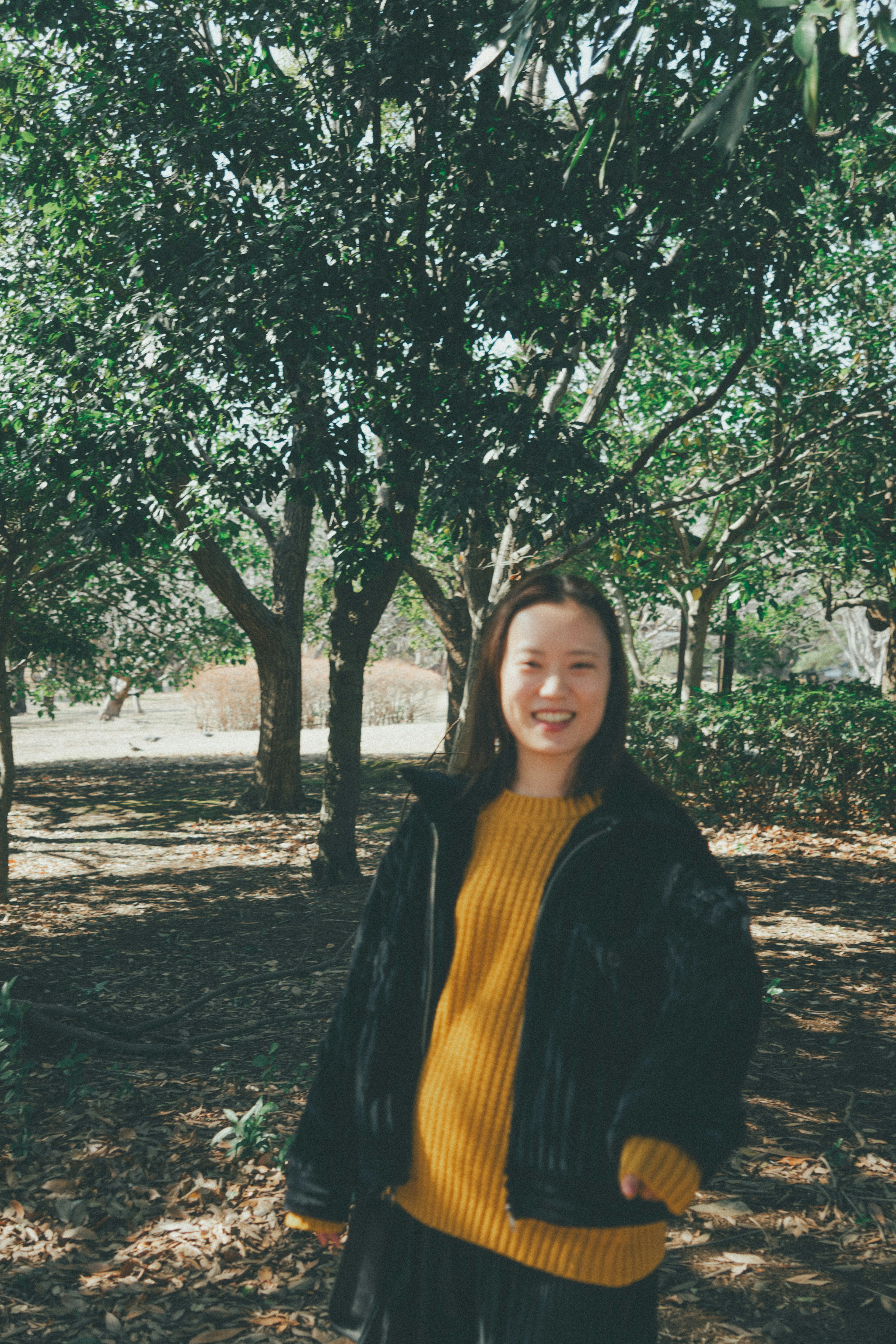 A woman standing among trees wearing a black jacket and yellow sweater with a smile