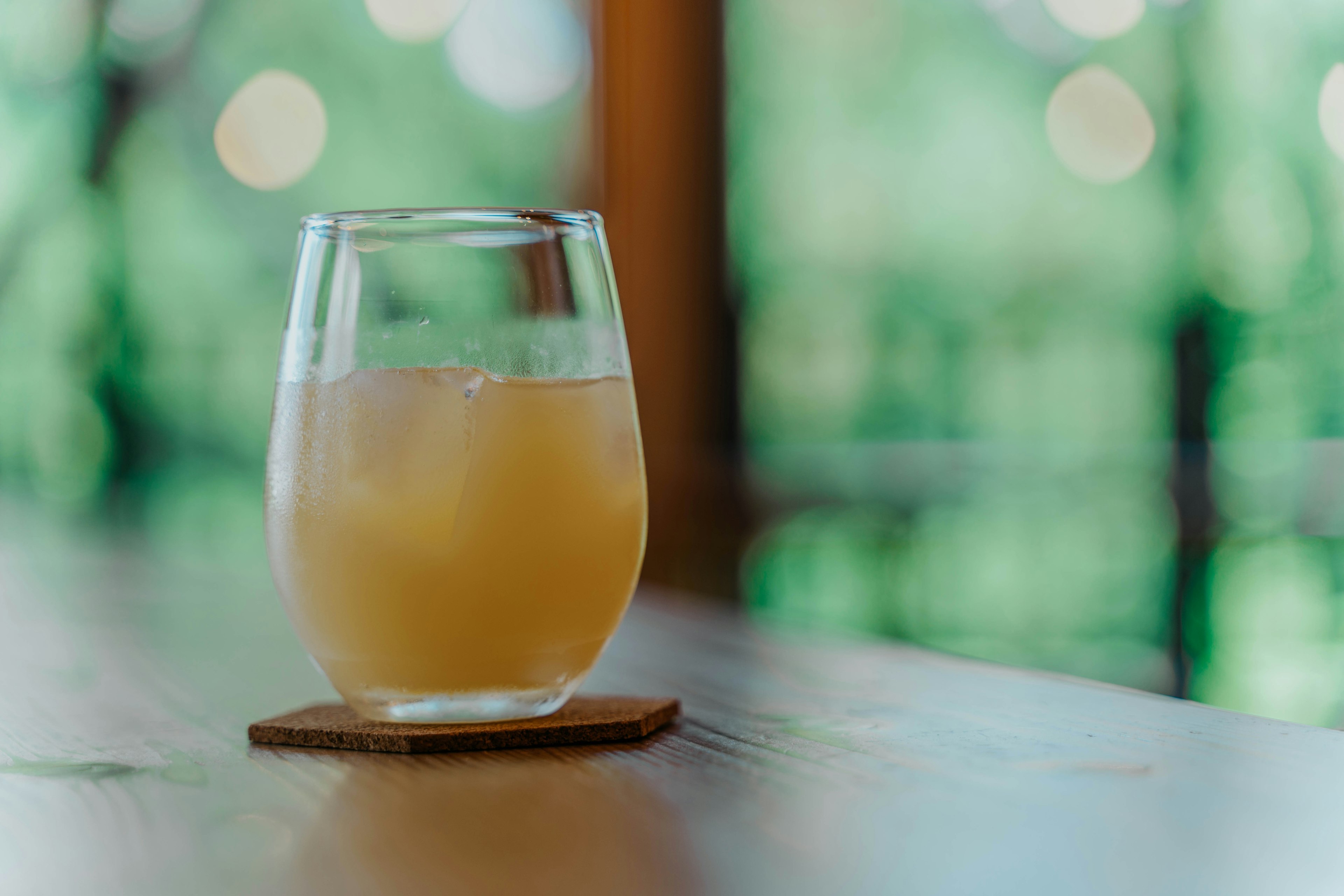 A transparent glass with a drink on a wooden coaster blurred green background