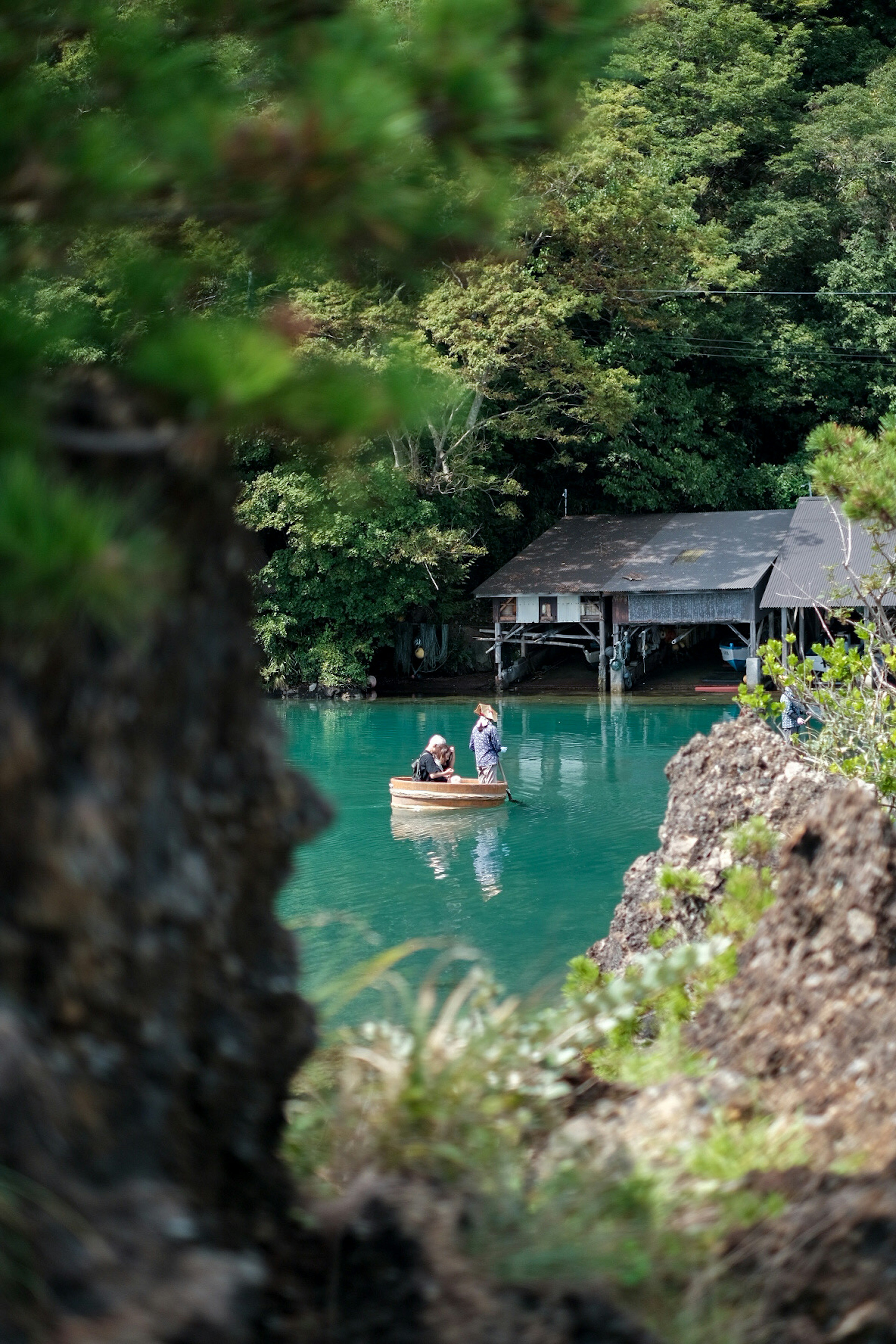 Two people in a boat on a blue lake surrounded by trees and a wooden hut