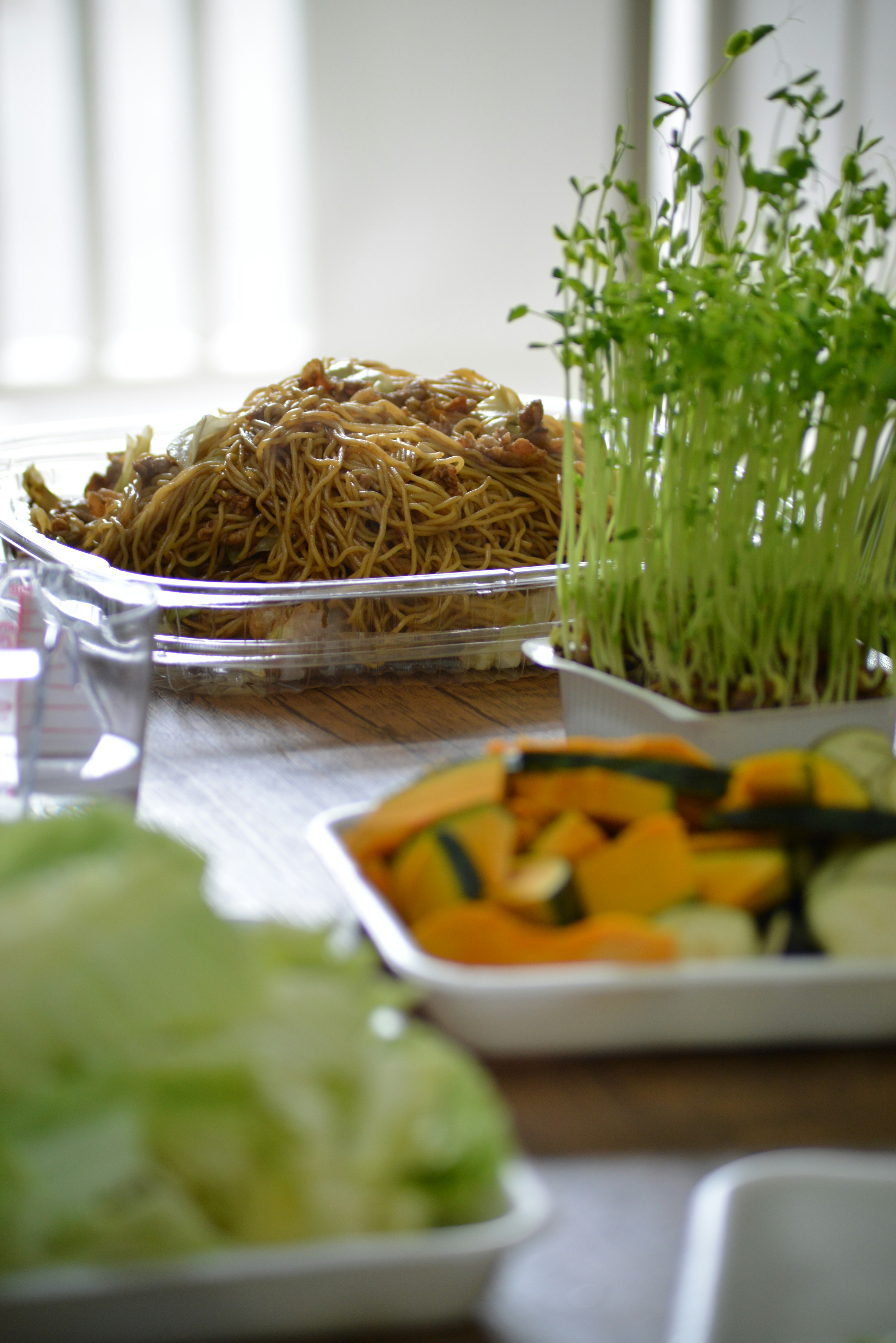 新鮮な野菜と豆苗が並ぶテーブルの上に、麺とカボチャの皿がある