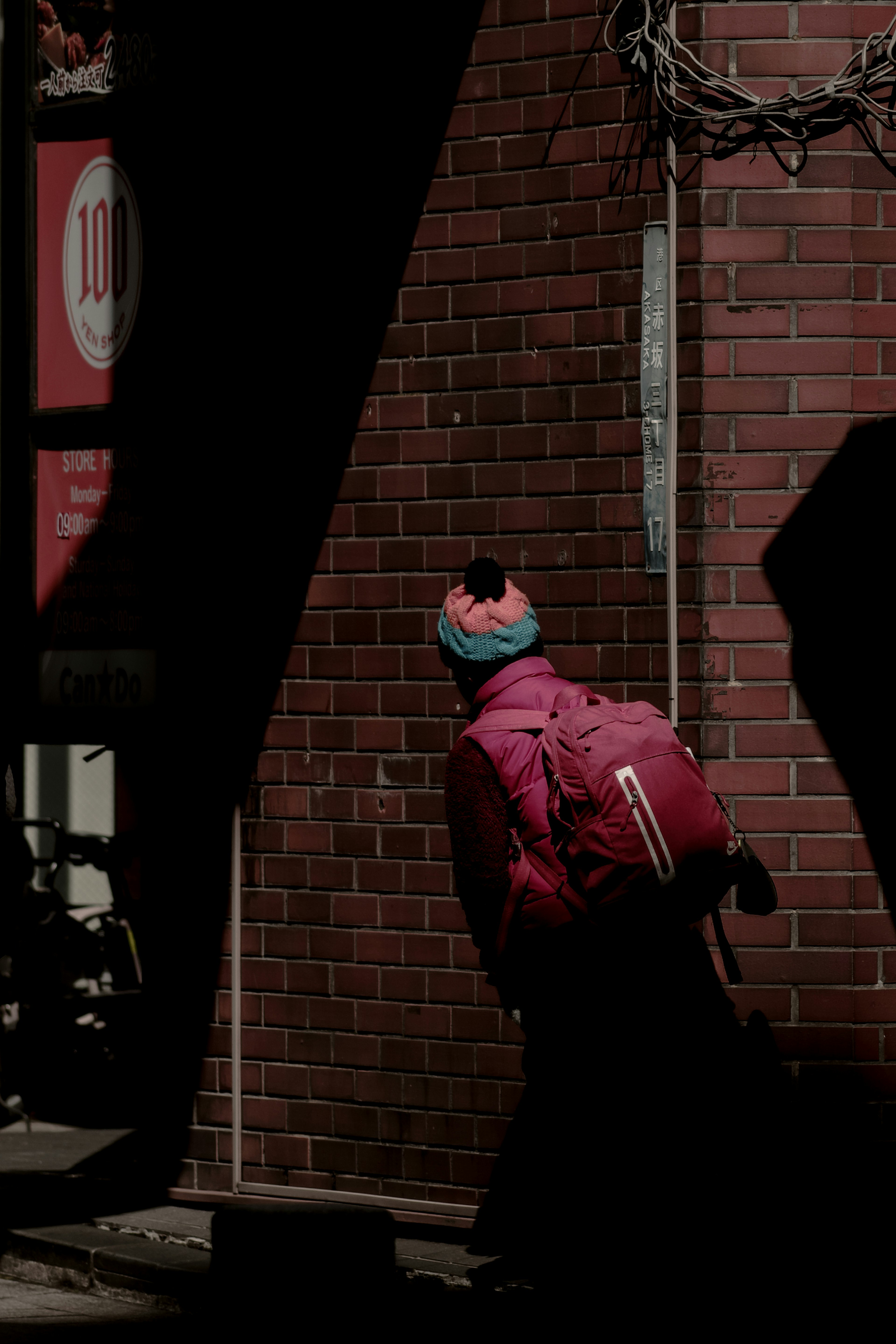 Une personne en veste rose et bonnet coloré s'appuie contre un mur en briques rouges avec un sac à dos