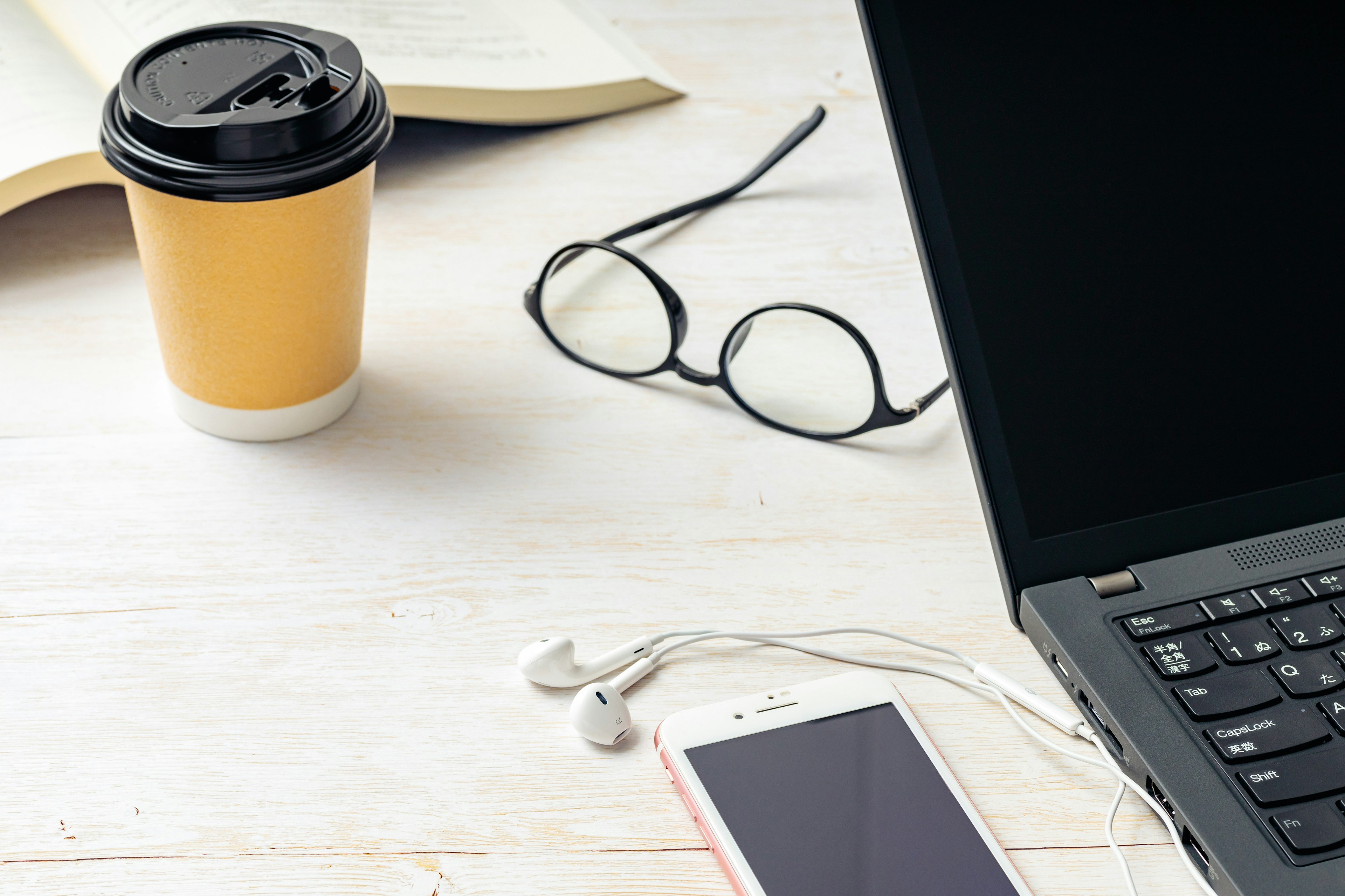 A laptop on a desk with a coffee cup nearby glasses and a smartphone with earphones