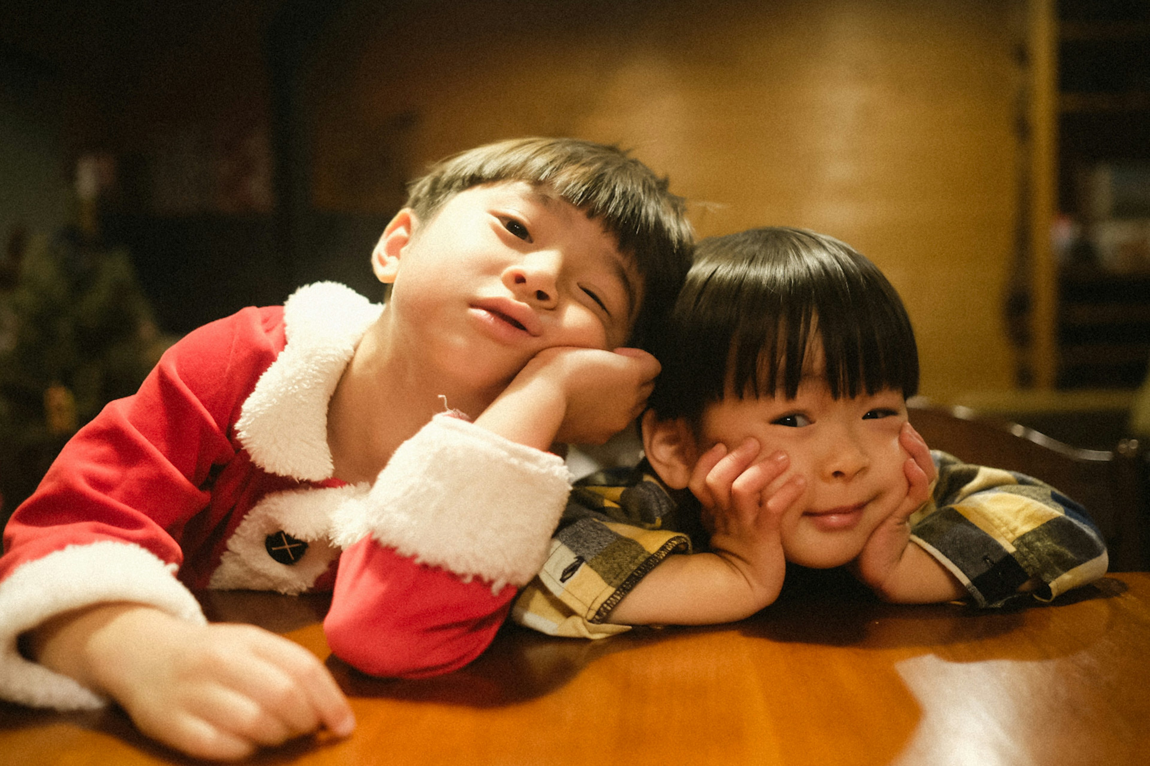 A boy in a Santa costume and a girl in a checkered shirt resting their heads on a table