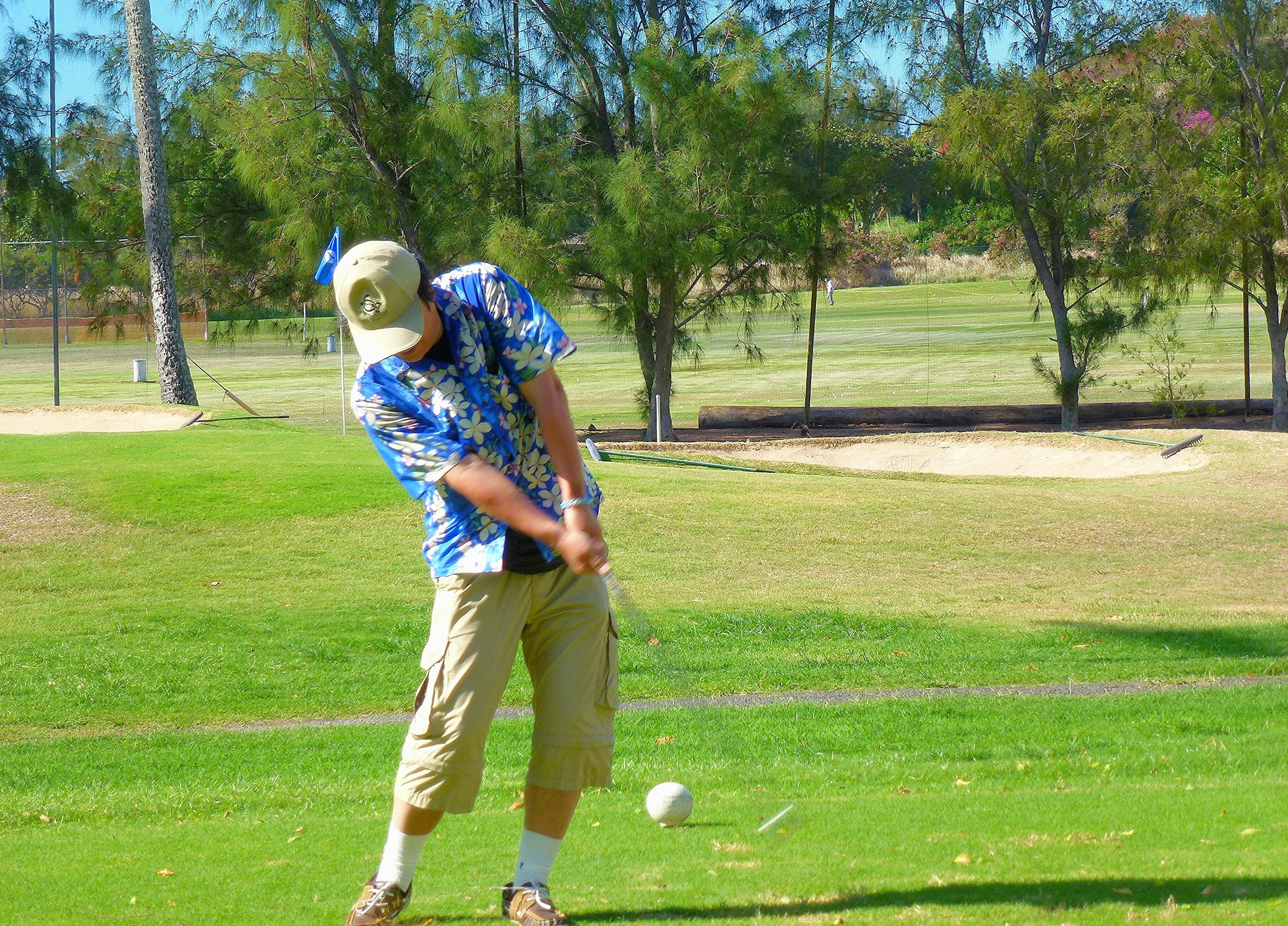 Mann spielt Golf in einem blauen Blumenhemd und Shorts in einem Golfschwung auf grünem Gras bei sonnigem Wetter