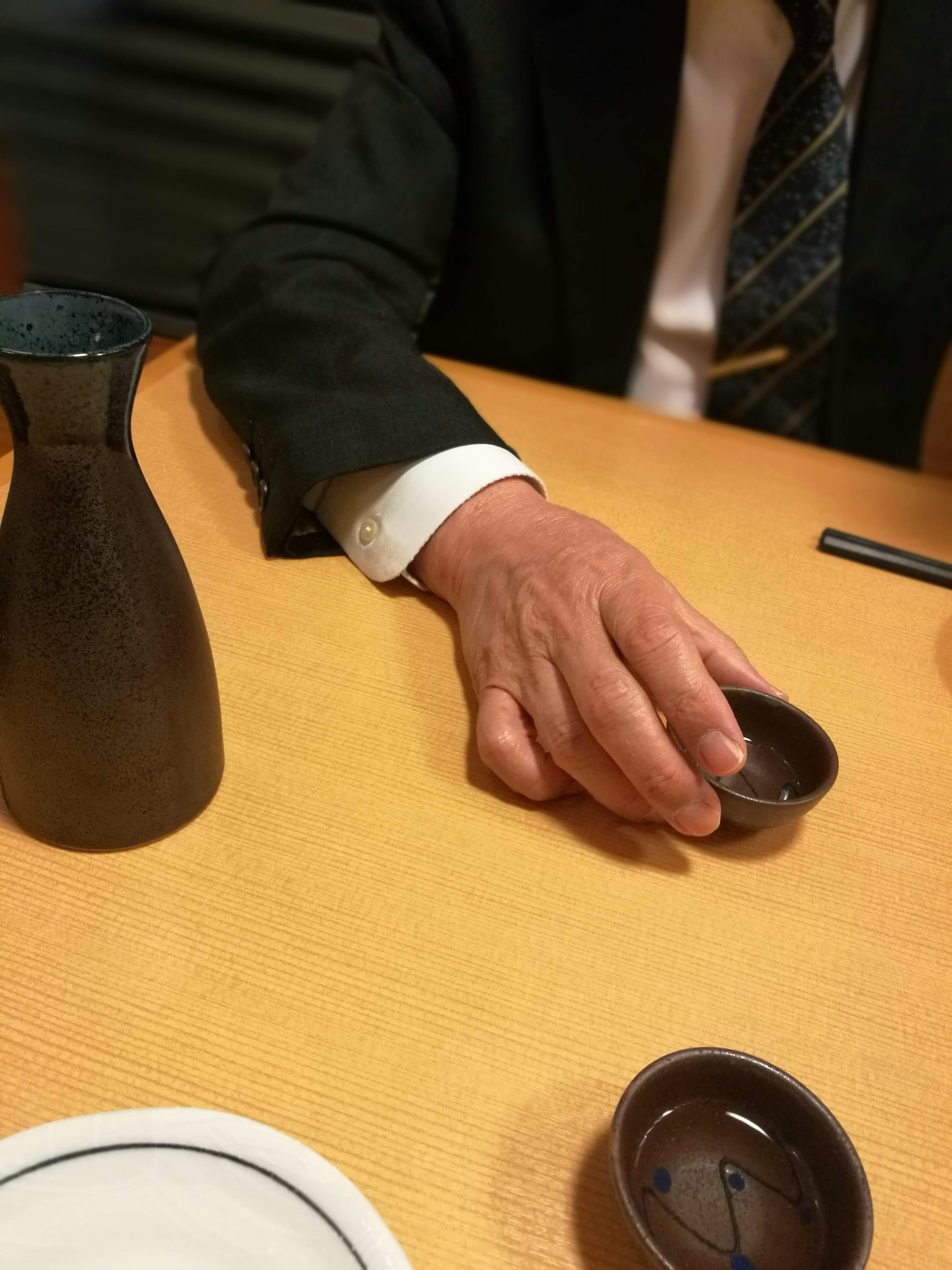 Die Hand eines Mannes, der eine Sake-Tasse neben einer Sake-Flasche auf einem Tisch hält