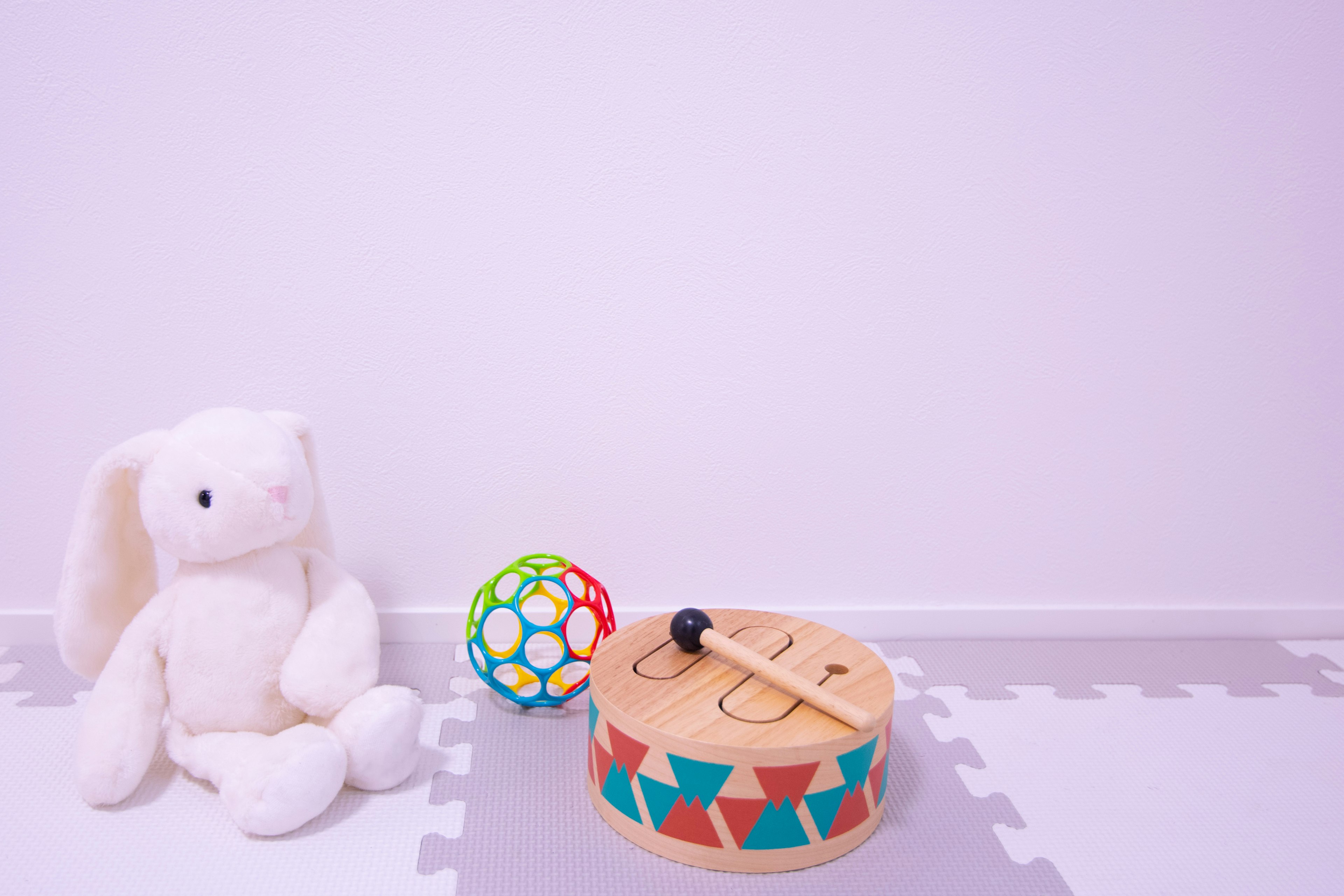 A plush white rabbit next to a colorful ball and a wooden drum on a white floor