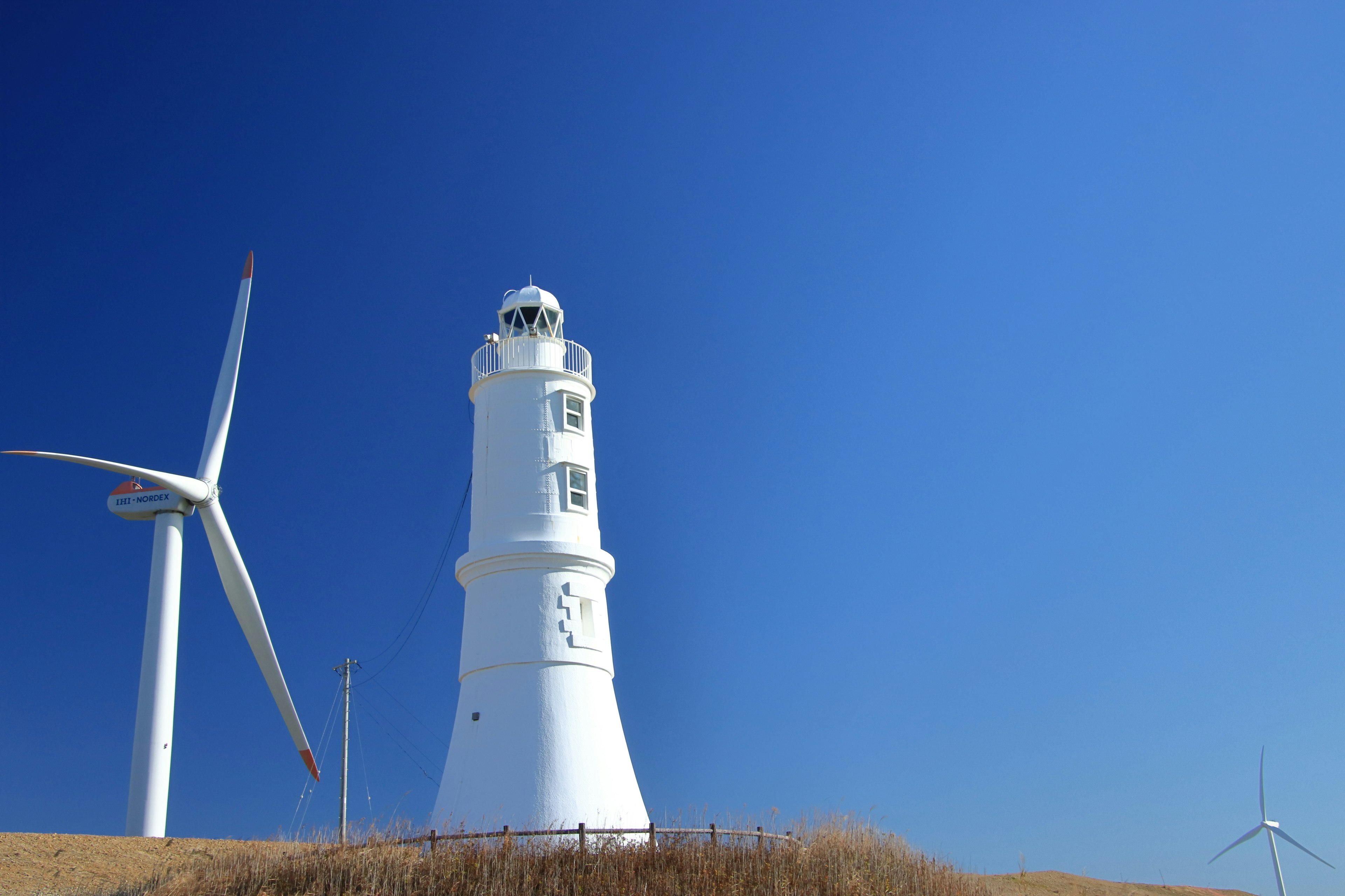 Faro bianco accanto a turbine eoliche sotto un cielo blu chiaro