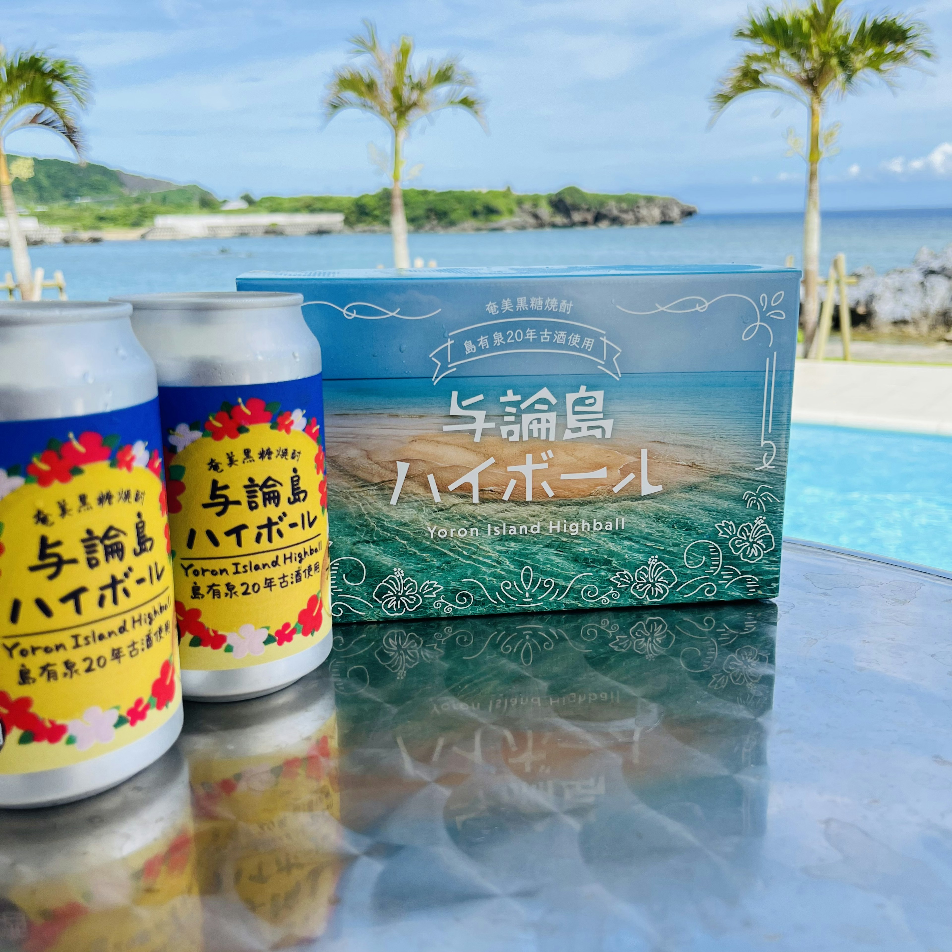 Cans of beer and a box placed on a table with a beach and palm trees in the background