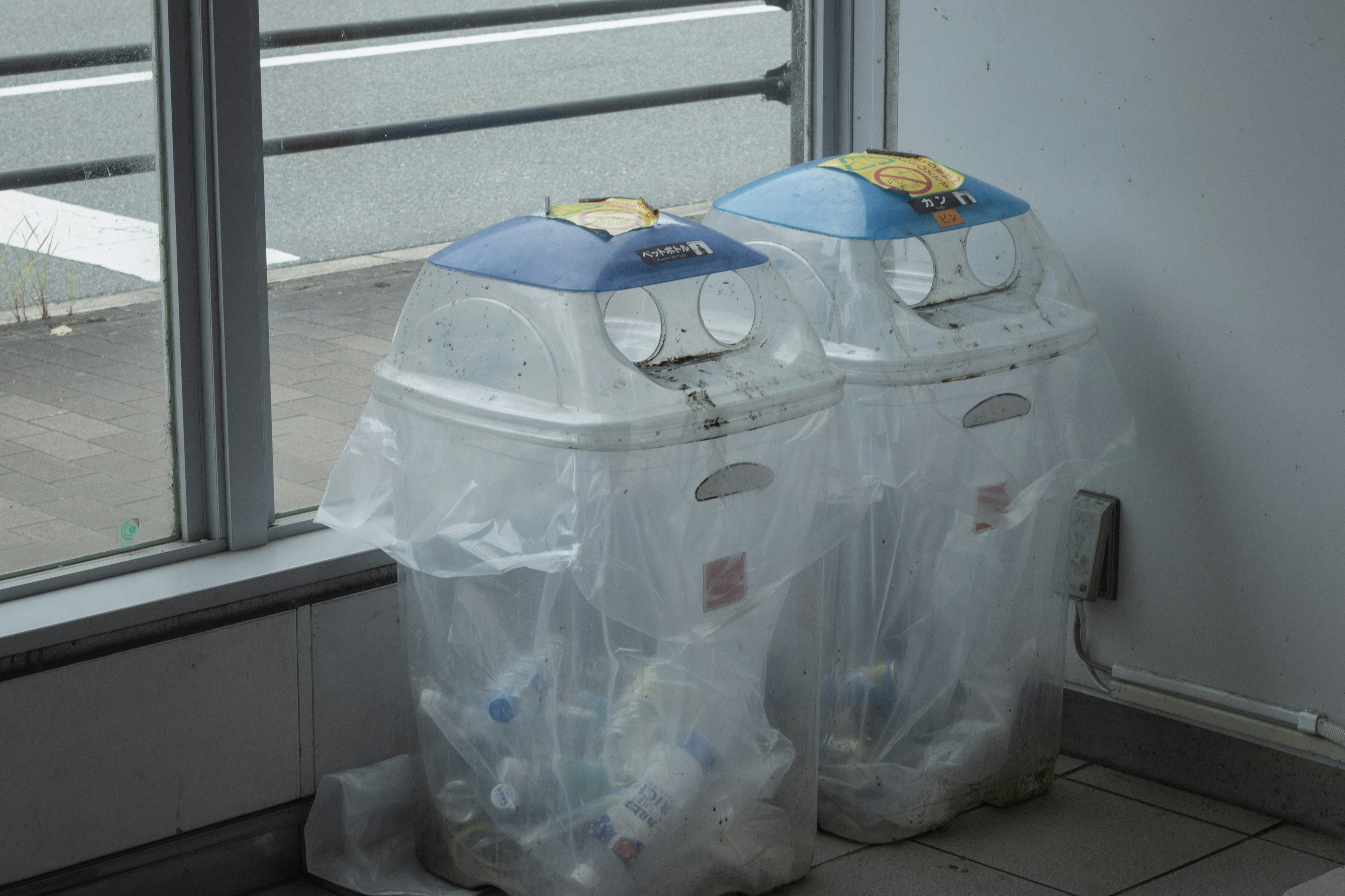 Two trash bins covered with clear bags near a window