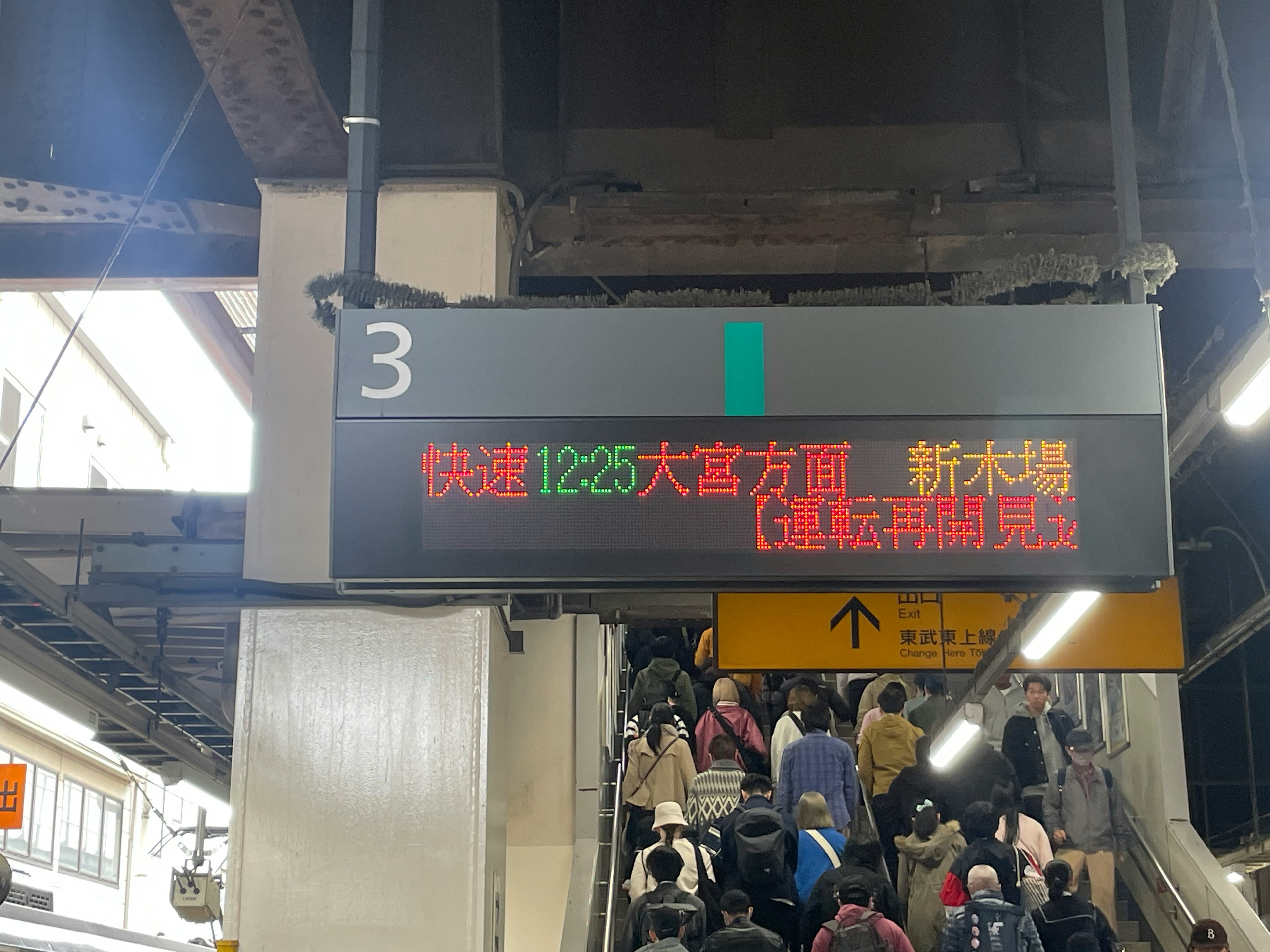 Panneau d'affichage numérique à la gare montrant les informations sur les trains avec des passagers sur les escalators